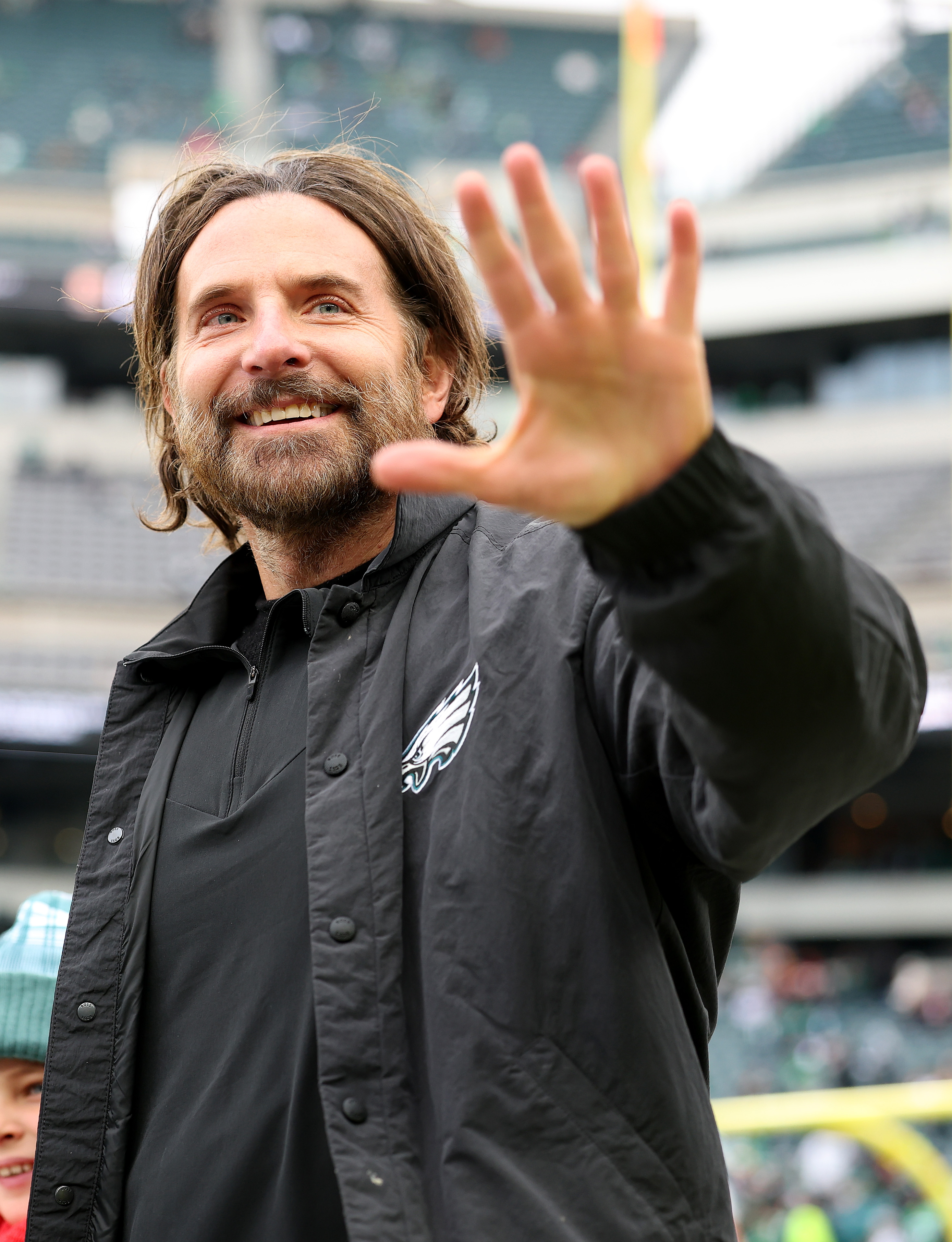 Cooper conveyed a warm expression with his eyes crinkling at the corners as he raised his hand in a wave, showcasing a full beard with prominent streaks of gray. This was taken during the NFC Championship Game at Lincoln Financial Field on January 26, 2025, in Philadelphia, Pennsylvania.