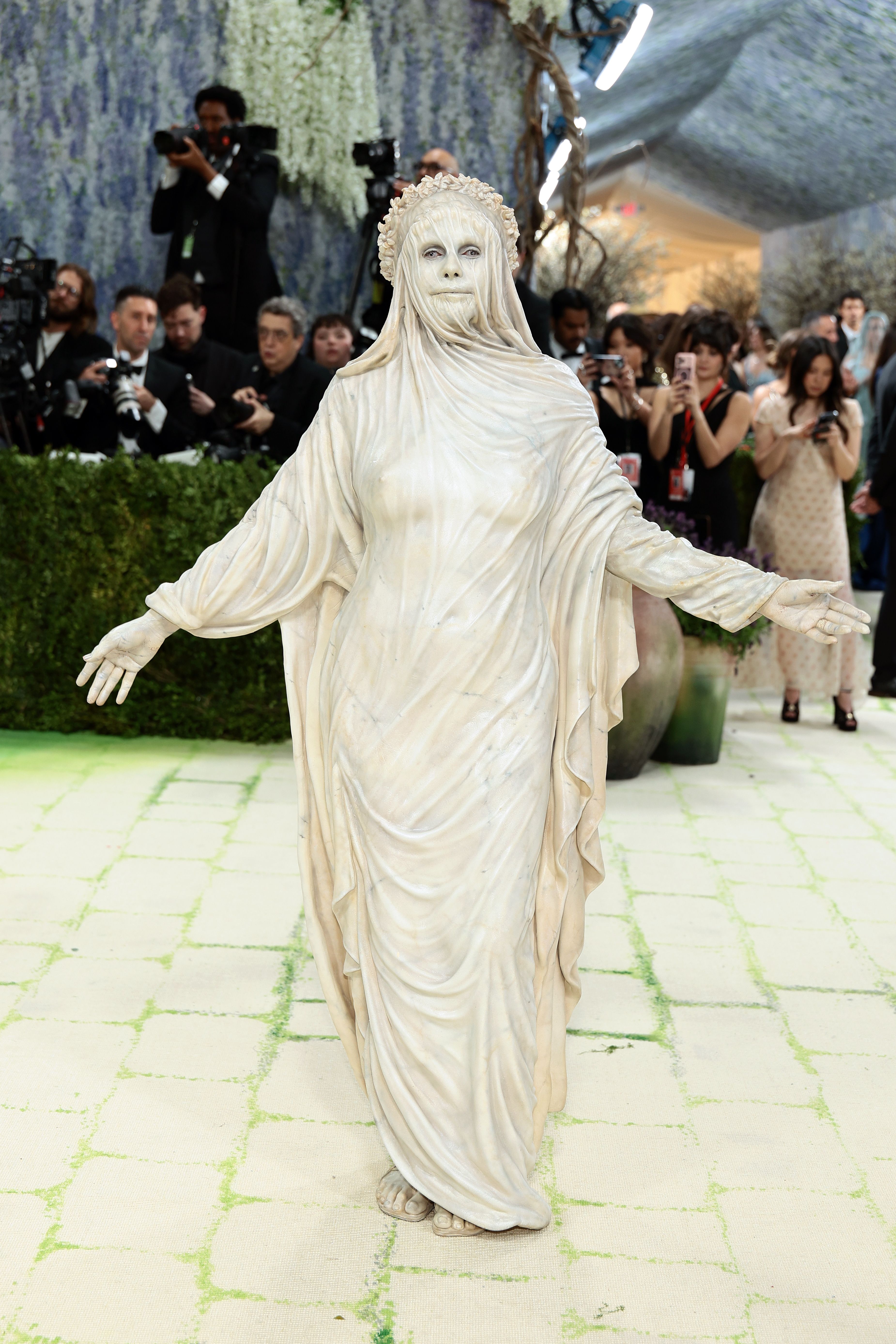 Heidi Klum stands arms outstretched on the carpet, her entire body — face, hands, and floor-length draped robes — painted ivory-white to replicate the texture and weight of classical marble, topped with a sculpted laurel wreath.