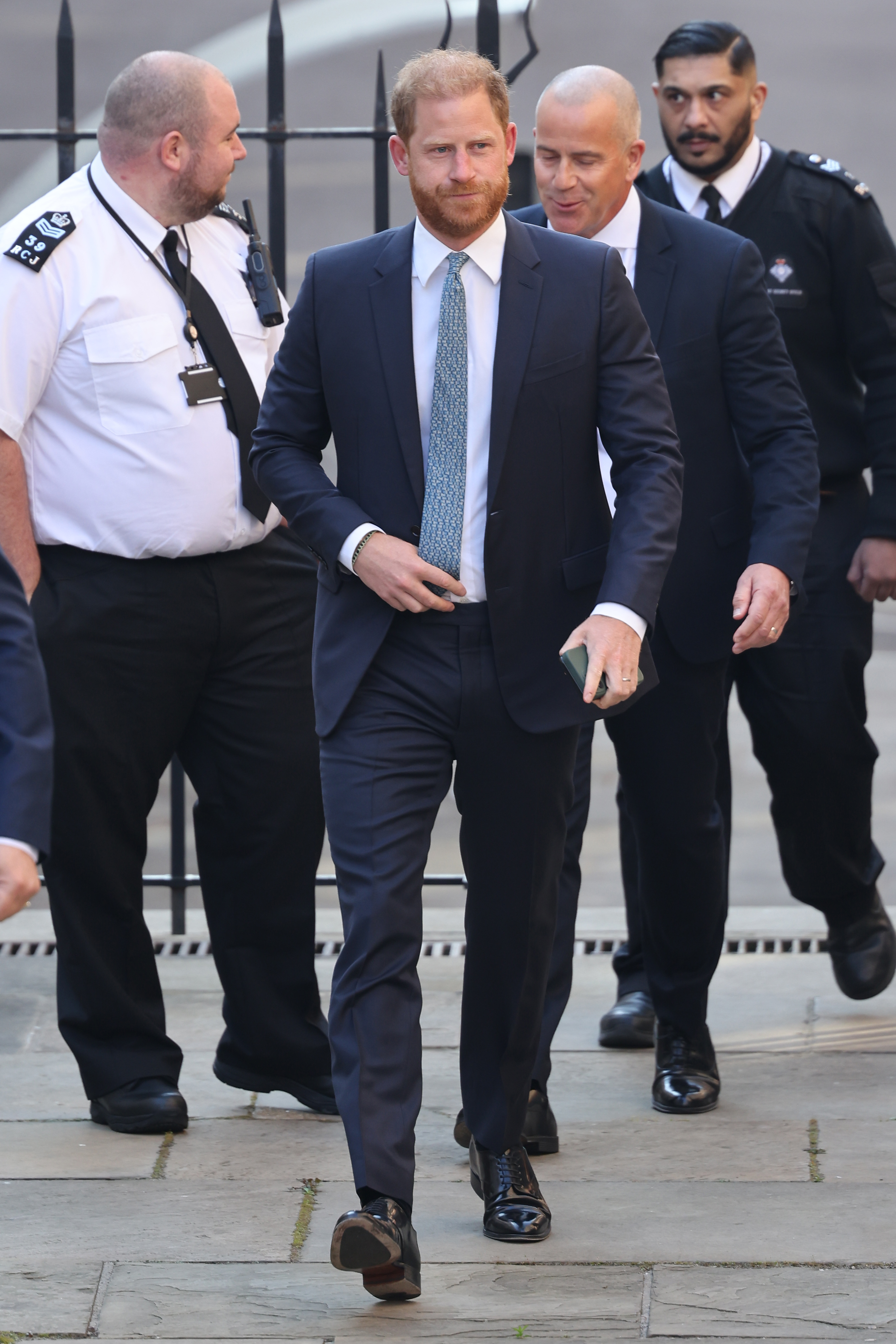 Prince Harry strides toward the Royal Courts of Justice surrounded by a discreet but visible security presence. Wearing a navy blue suit and a patterned tie, he looks composed yet determined — his gaze focused ahead.
