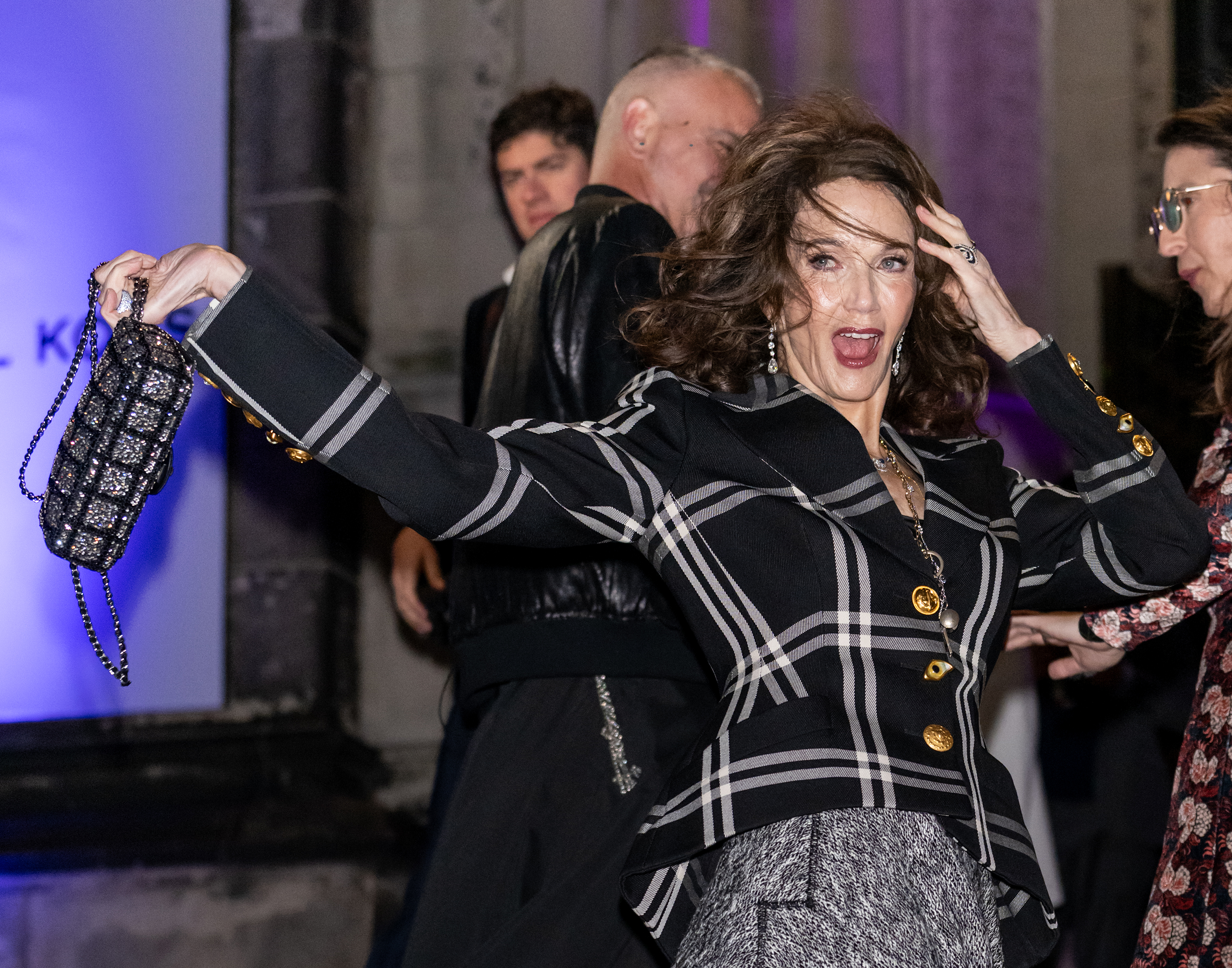 Lynda Carter strikes a playful pose outside the Michael Kors event, tossing her hair and flashing a sparkling handbag. Dressed in her bold plaid blazer with gold buttons and a textured gray skirt, she exudes energy and charisma while delighting photographers with her animated flair.