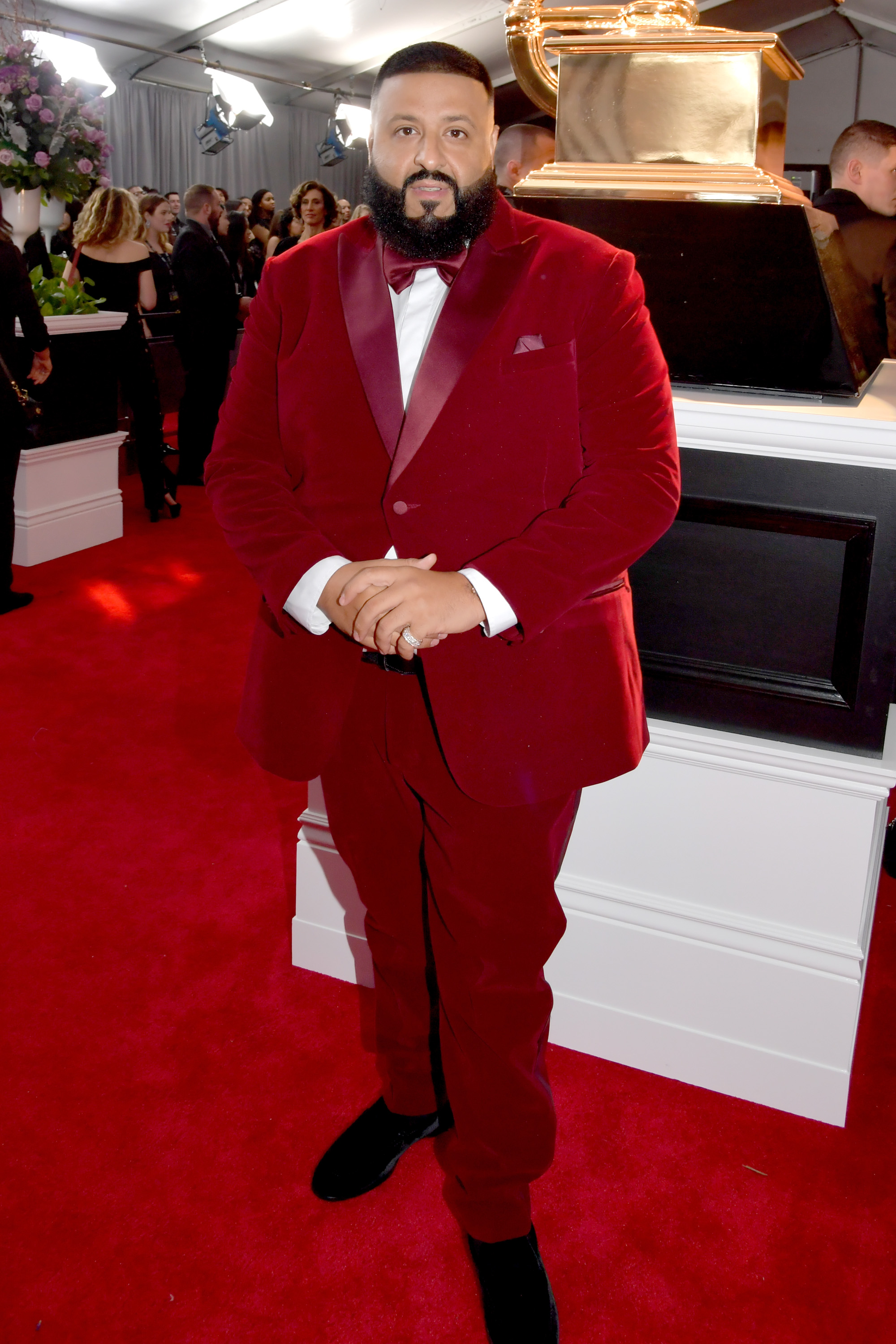 On the Grammy Awards red carpet, DJ Khaled arrives in a rich red velvet tuxedo complete with a satin bow tie and sleek black shoes, posing in front of the iconic golden gramophone display beneath bright event lighting and a flurry of activity behind him.