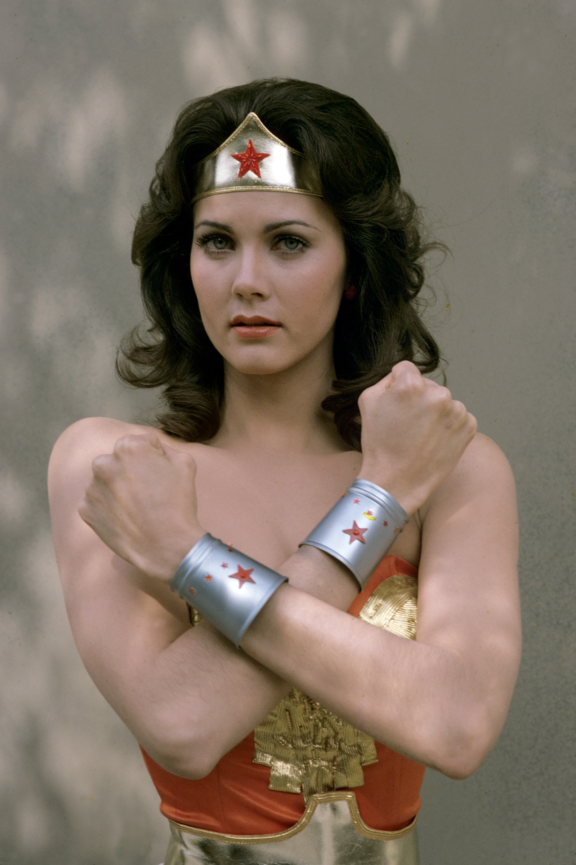 Lynda Carter, in full "Wonder Woman" costume, strikes the iconic power pose during a promotional shoot for the television series. Wearing her signature gold tiara and silver bracelets adorned with red stars, she gazes into the camera with quiet intensity.