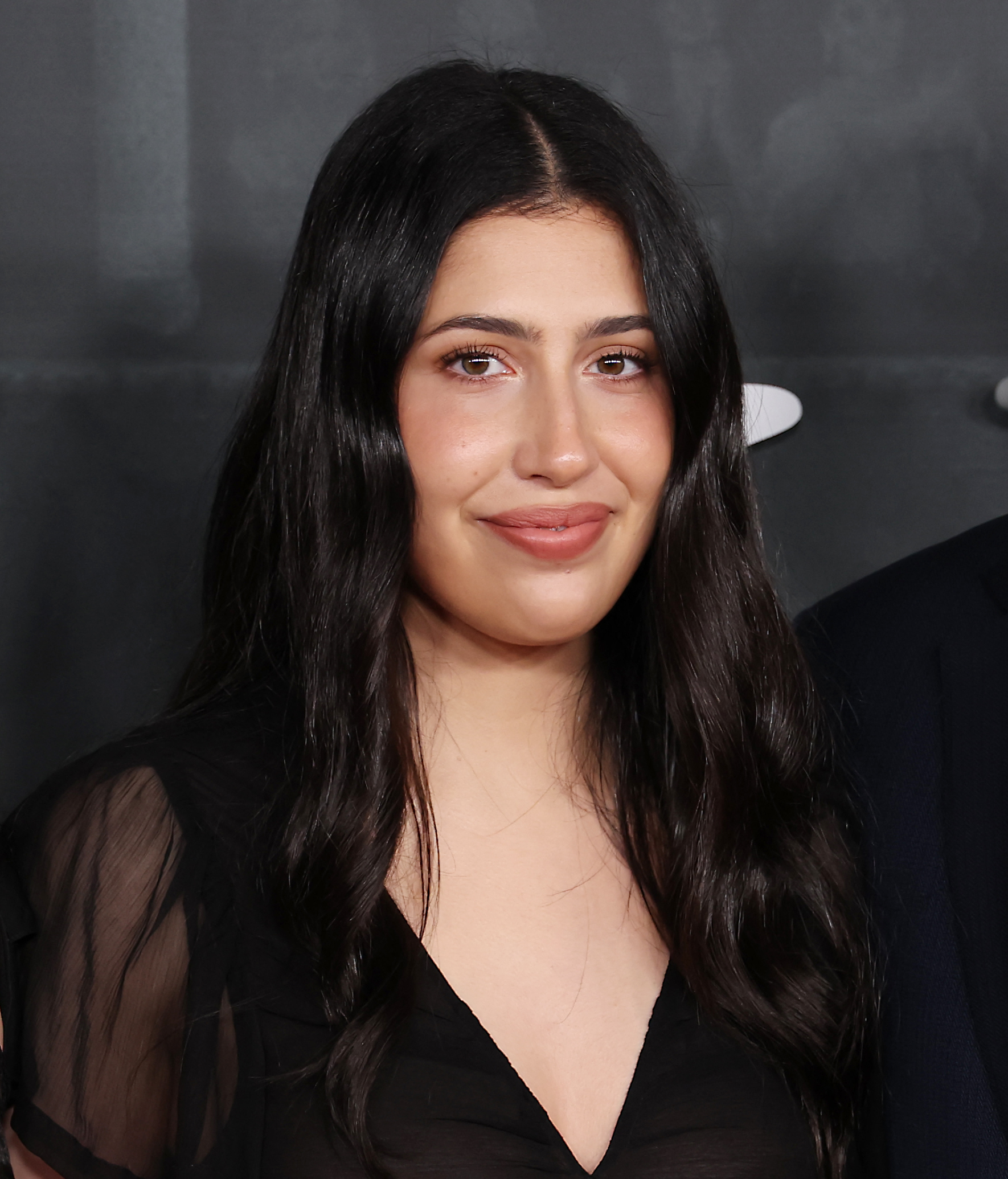 As the Sandlers pose together, one photographer zooms in to capture Sadie Sandler at the "Jay Kelly" premiere. She looks radiant, her black hair parted in the middle and flowing to her shoulders, complemented by subtle, flawless makeup.