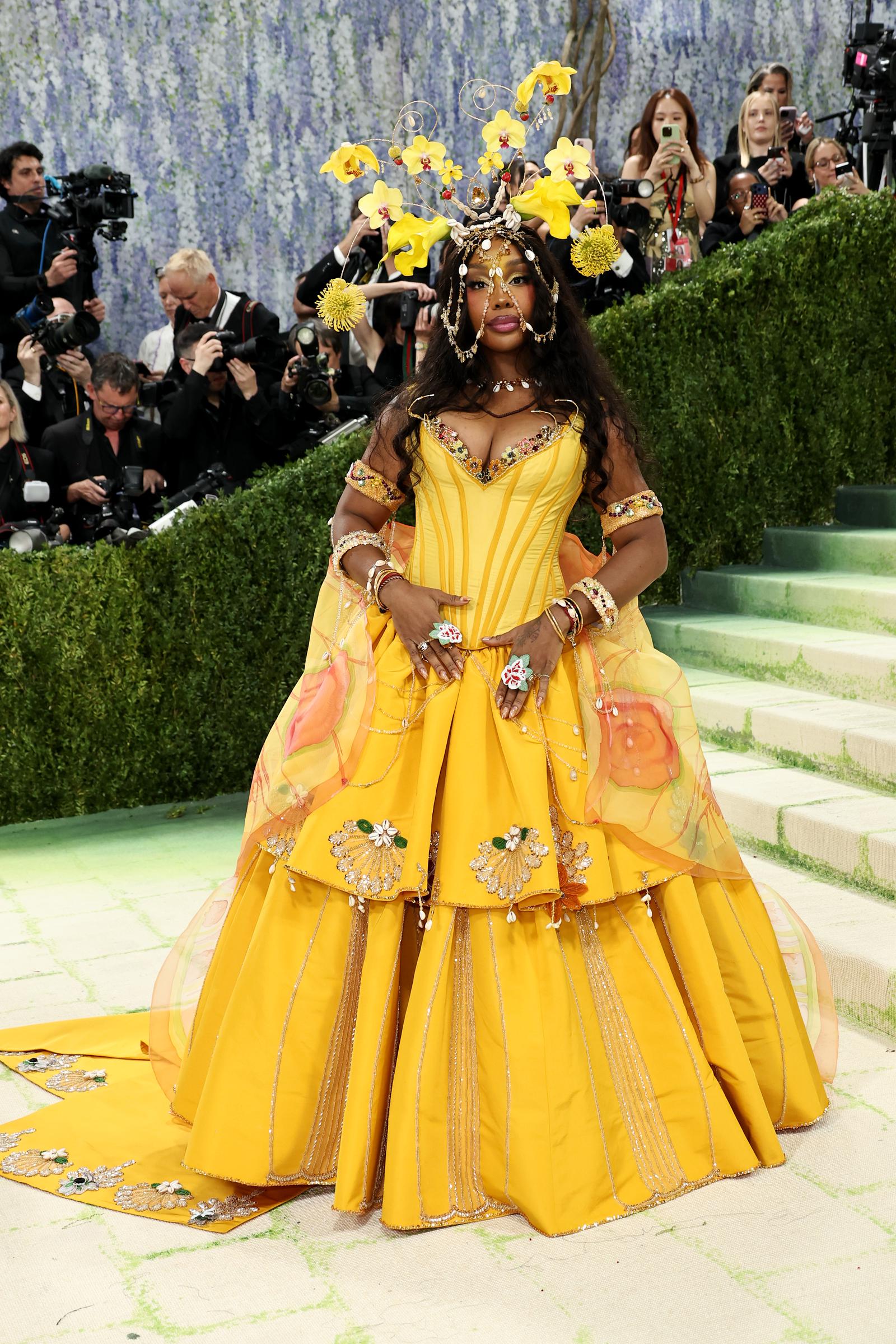 SZA stuns in a voluminous golden yellow tiered ballgown with floral embroidery, sheer organza panels with painted blooms, a jewelled corset bodice, stacked cocktail rings and bracelets, and a towering headdress of yellow orchids, wire spirals, and shell embellishments cascading over her face.