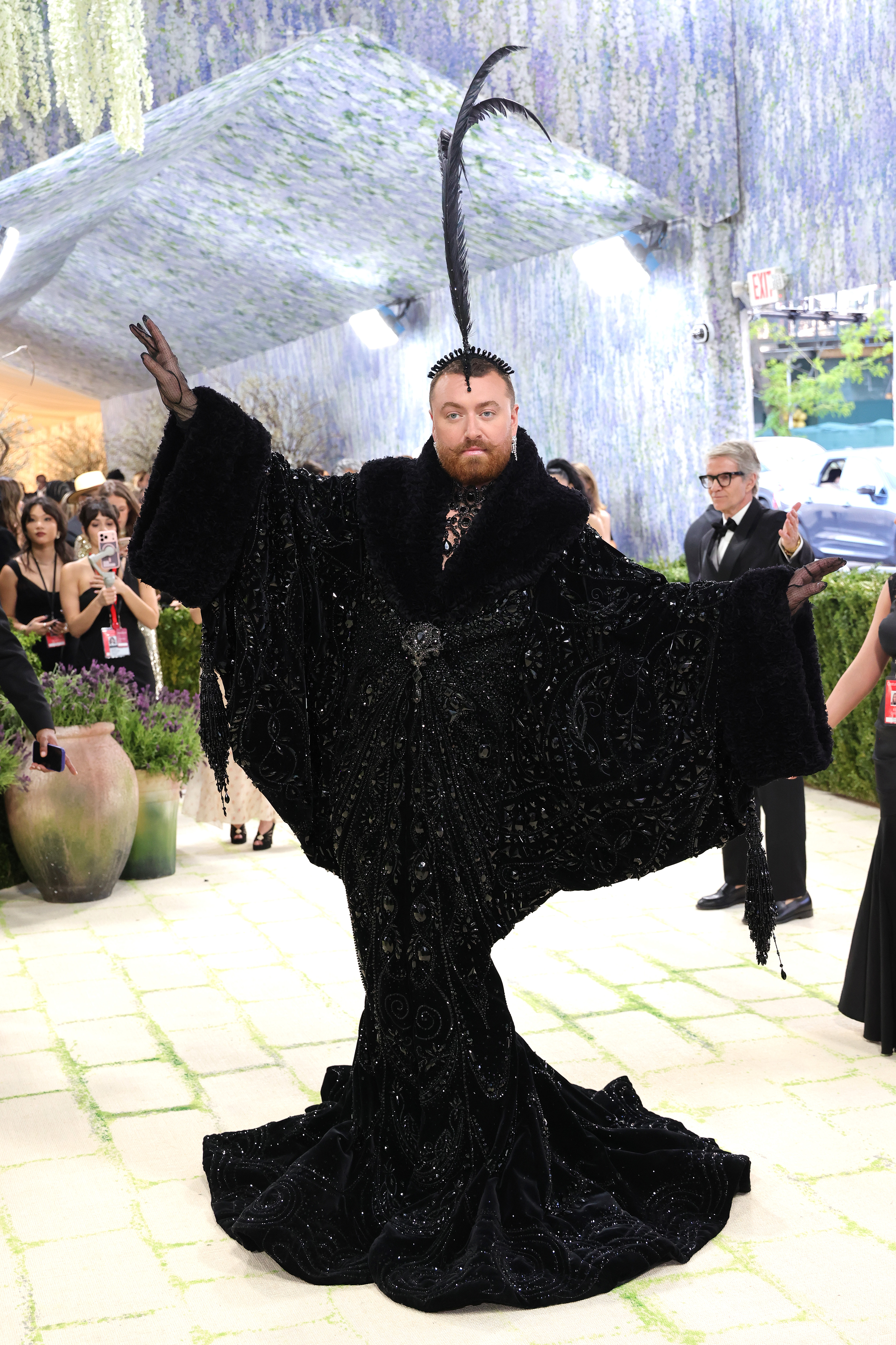 A wider shot of Sam Smith arms outstretched, showcasing the full dramatic wingspan of the black beaded robe, its fur-trimmed collar, trailing sequinned train, sheer black gloves, beaded tassel cuffs, and a single soaring black plume headpiece against the open-air backdrop.