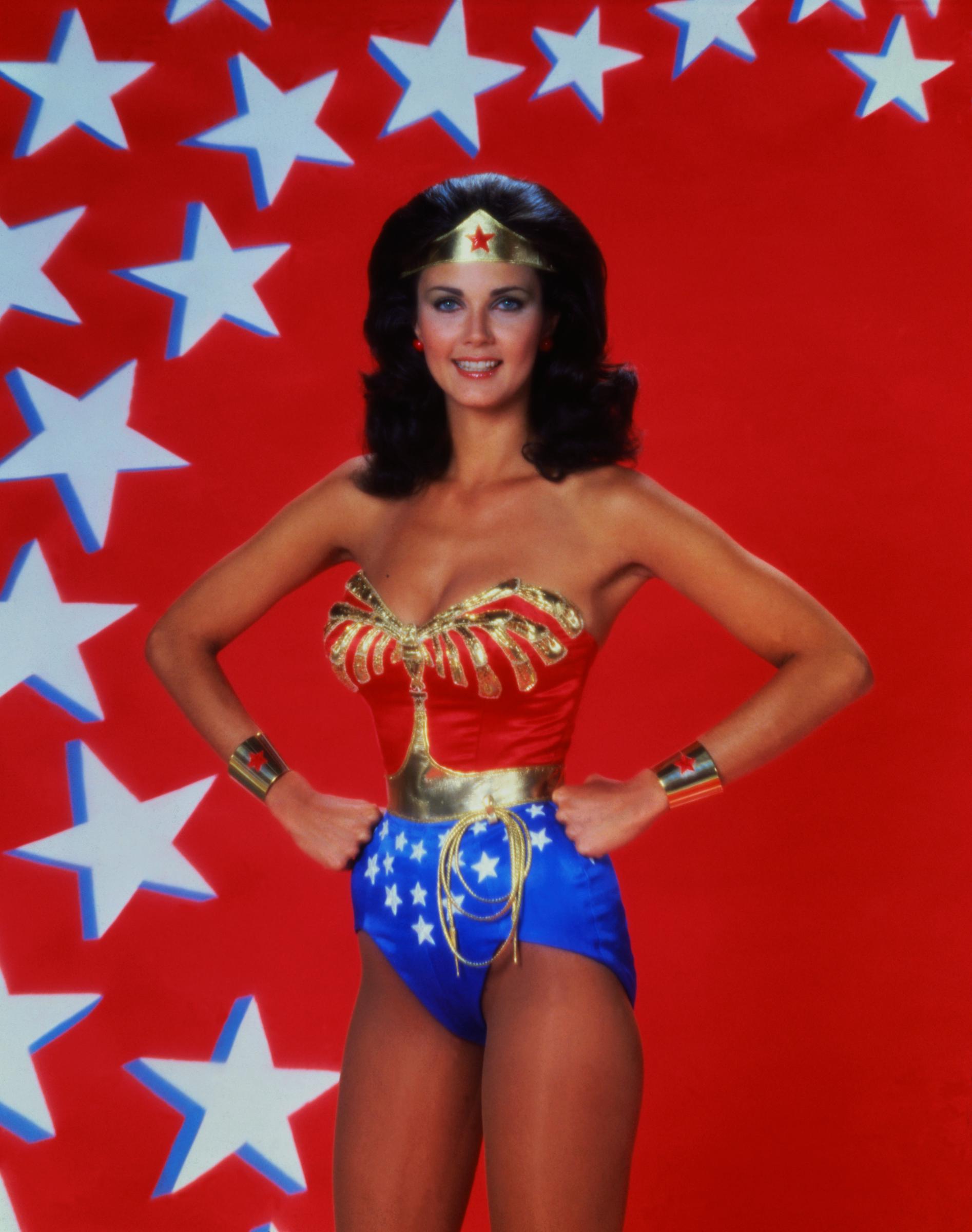 Lynda Carter stands confidently in her classic "Wonder Woman" costume against a bold red backdrop scattered with white stars. Hands on hips, she flashes a bright smile while wearing the updated version of the superhero’s iconic outfit—featuring a gold eagle bodice, blue star-spangled shorts, and her signature tiara.