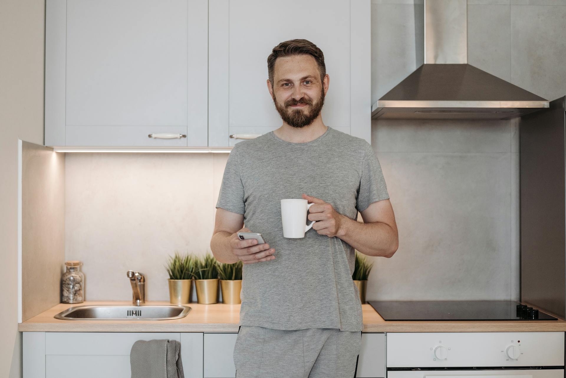 A man holding a phone and a mug while standing in the kitchen | Source: Pexels