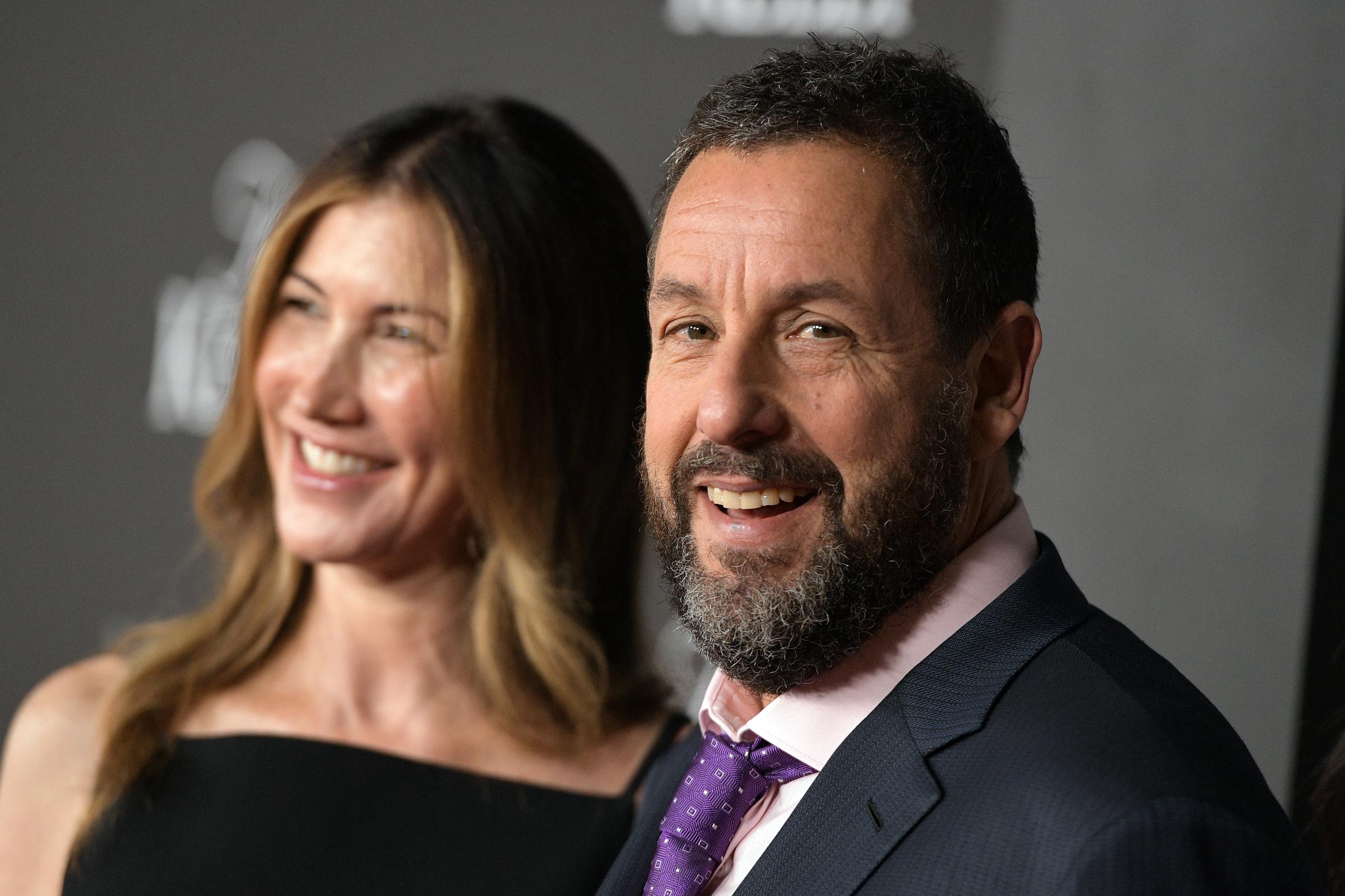 Adam Sandler is joined by his wife, Jackie Sandler, on the red carpet at Netflix’s "Jay Kelly" premiere. Adam beams with a broad smile, while his wife smiles subtly, slightly out of focus in the background.