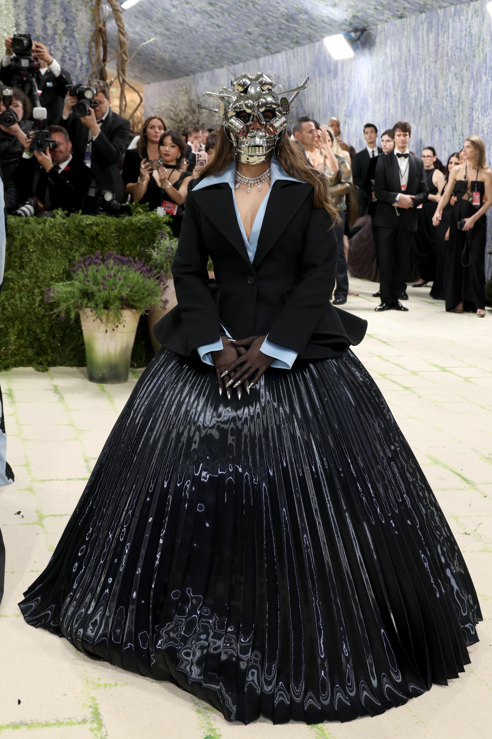 Ananya Birla in a black structured blazer with powder blue lapels and cuffs over a voluminous black pleated iridescent ballgown skirt with a swirling silver marbled pattern, silver pointed nail tips, a diamond choker, and a menacing chrome skull mask with protruding spikes covering her entire face.
