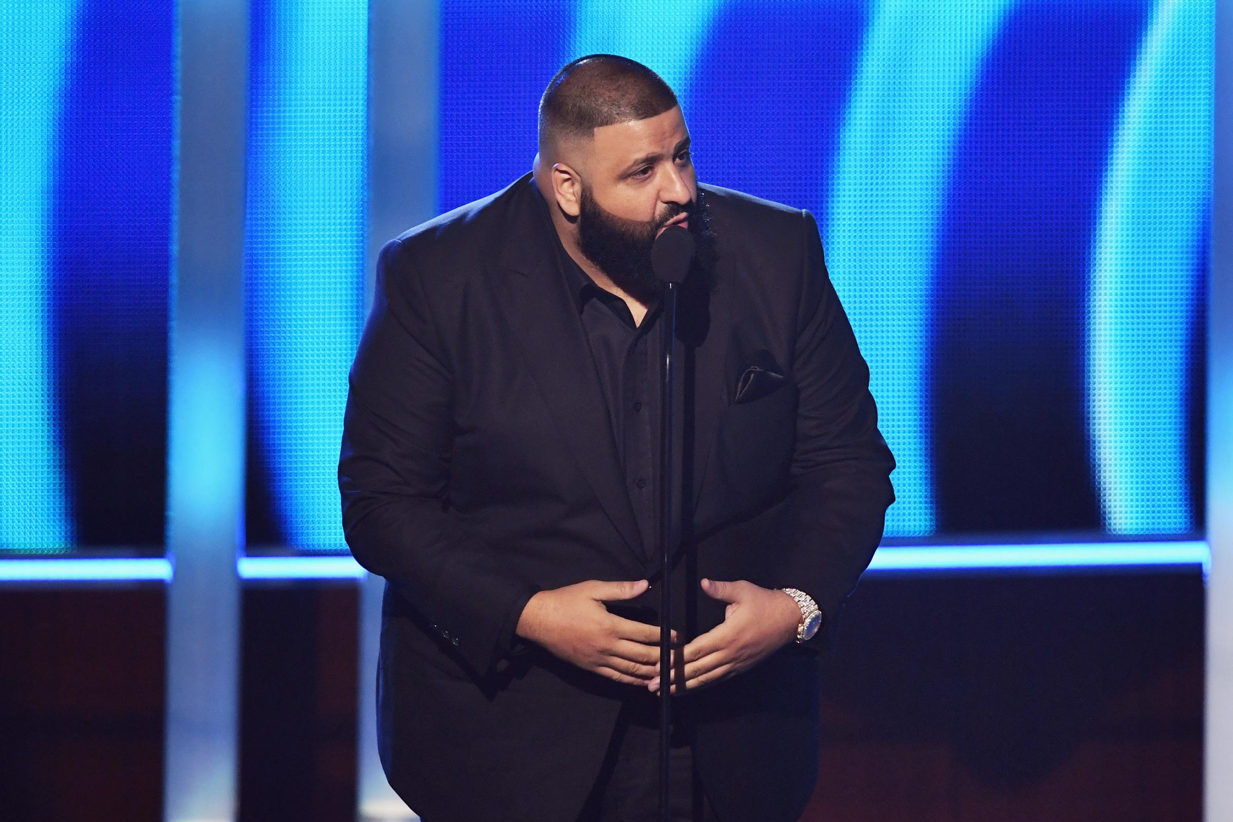 Dressed in an all-black suit, DJ Khaled leans toward the microphone mid-speech onstage, illuminated by cool-toned lighting that casts a subtle glow across his neatly groomed beard and tailored silhouette during a live award show appearance.