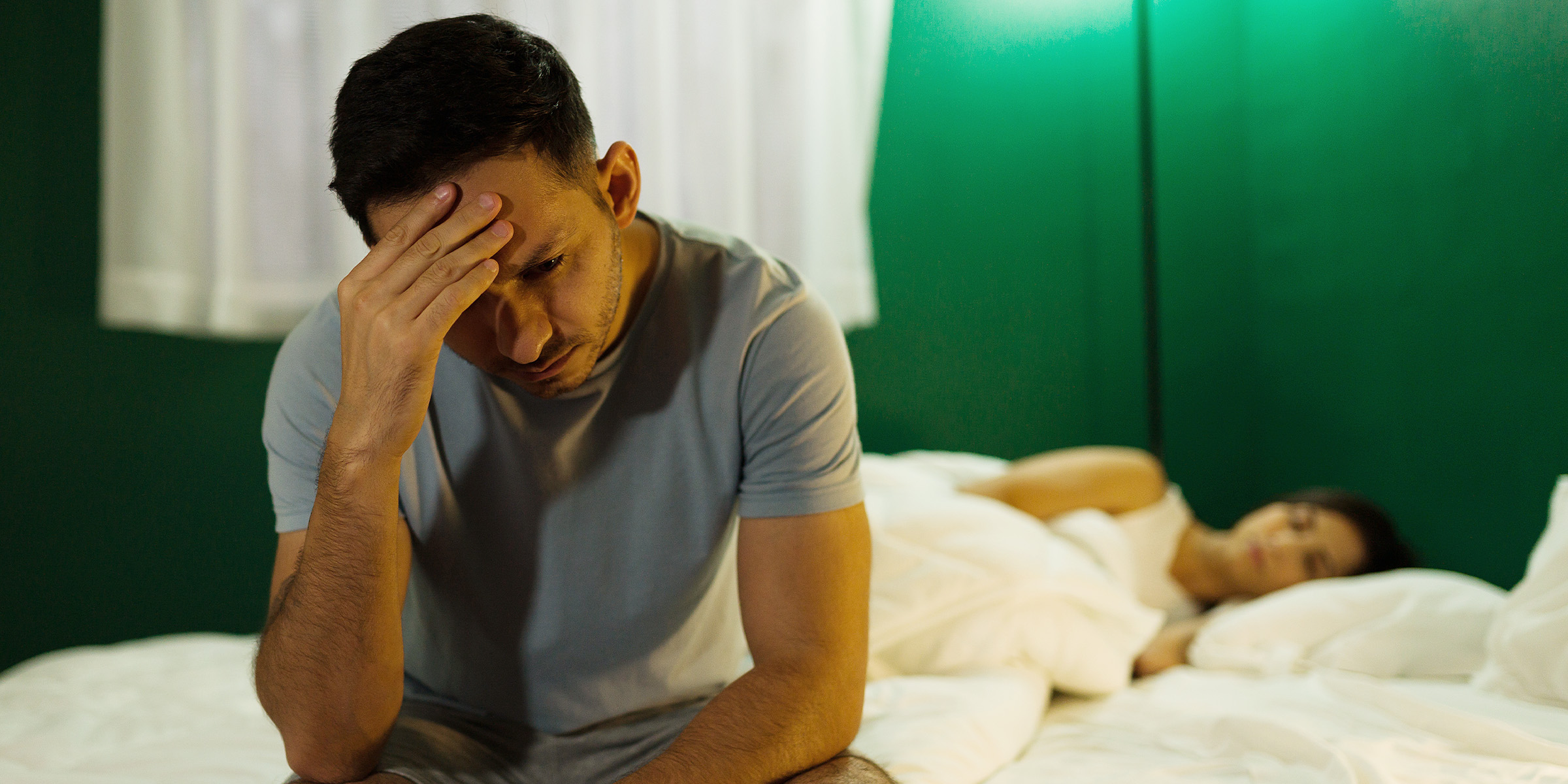 A stressed husband sitting on a bed while his wife sleeps | Source: Shutterstock