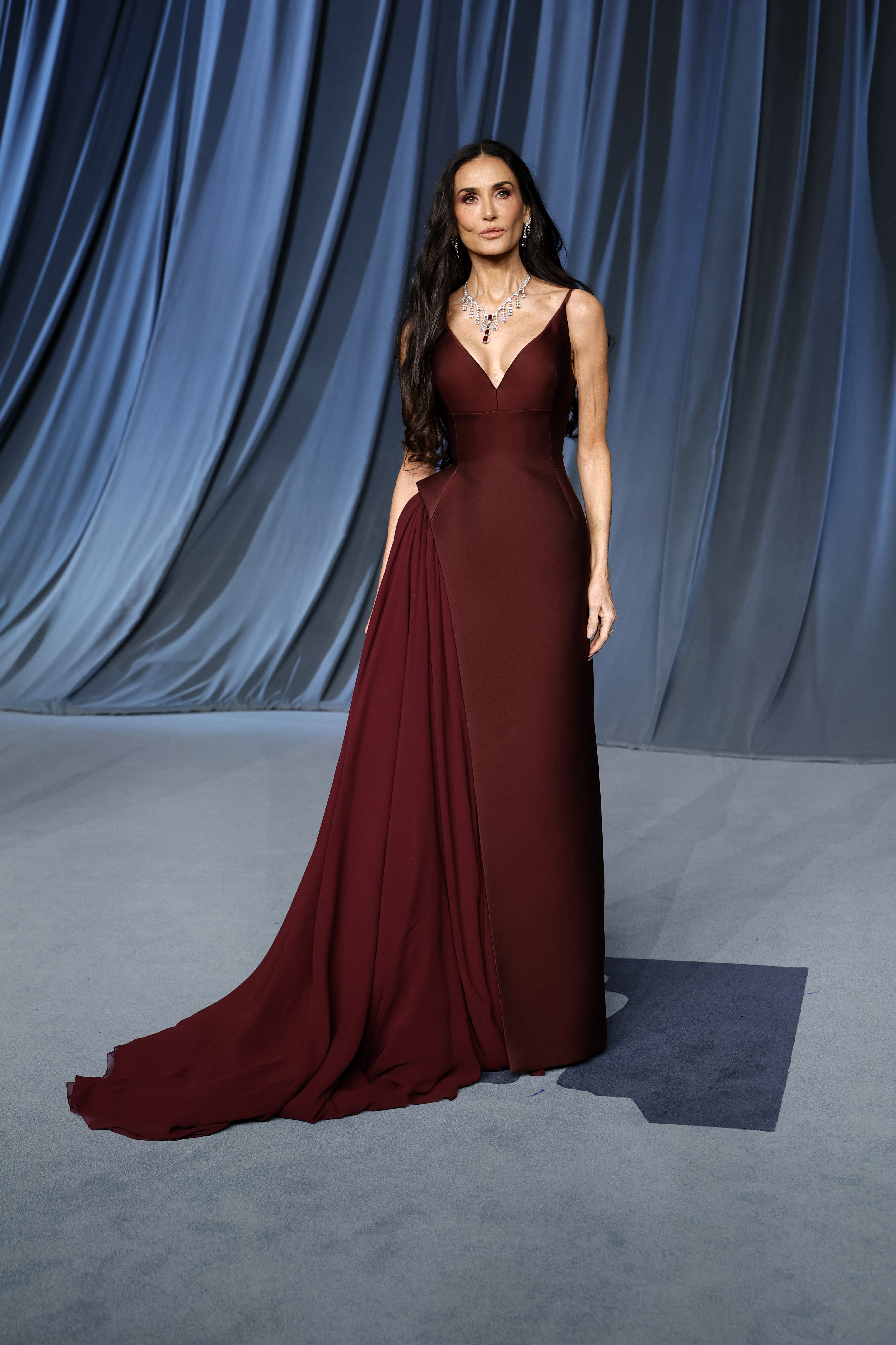 Demi Moore stands tall with a composed gaze straight ahead, her arms gently at her sides as the rich maroon gown pools around her feet. The flawless fit captures the full impact of her red carpet arrival moment.