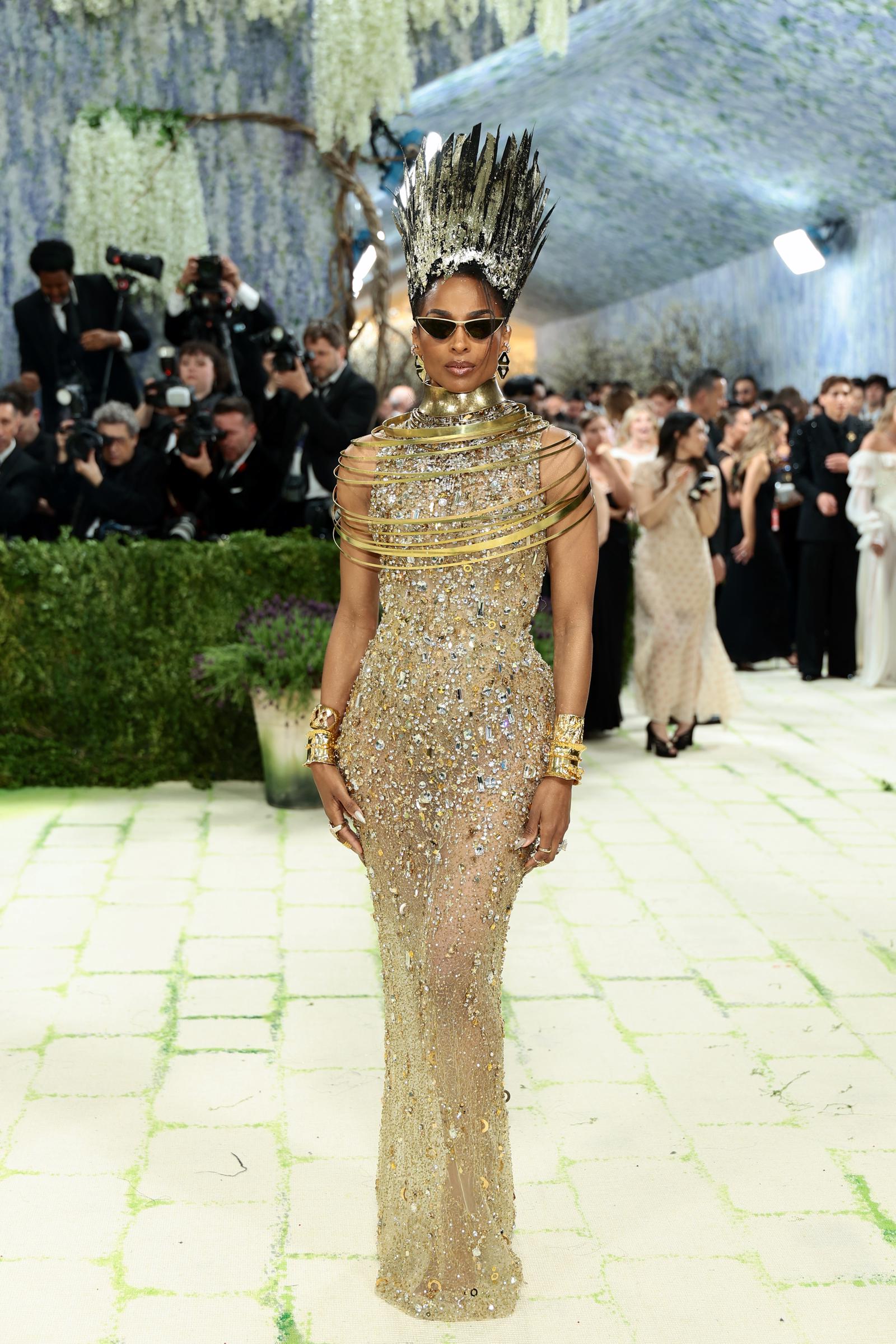 Ciara in a sheer crystal and gold embellished column gown with a structured multi-ring gold shoulder collar, thick gold cuff bracelets, cat-eye sunglasses, drop earrings, and a towering black gilded spiked crown headdress.