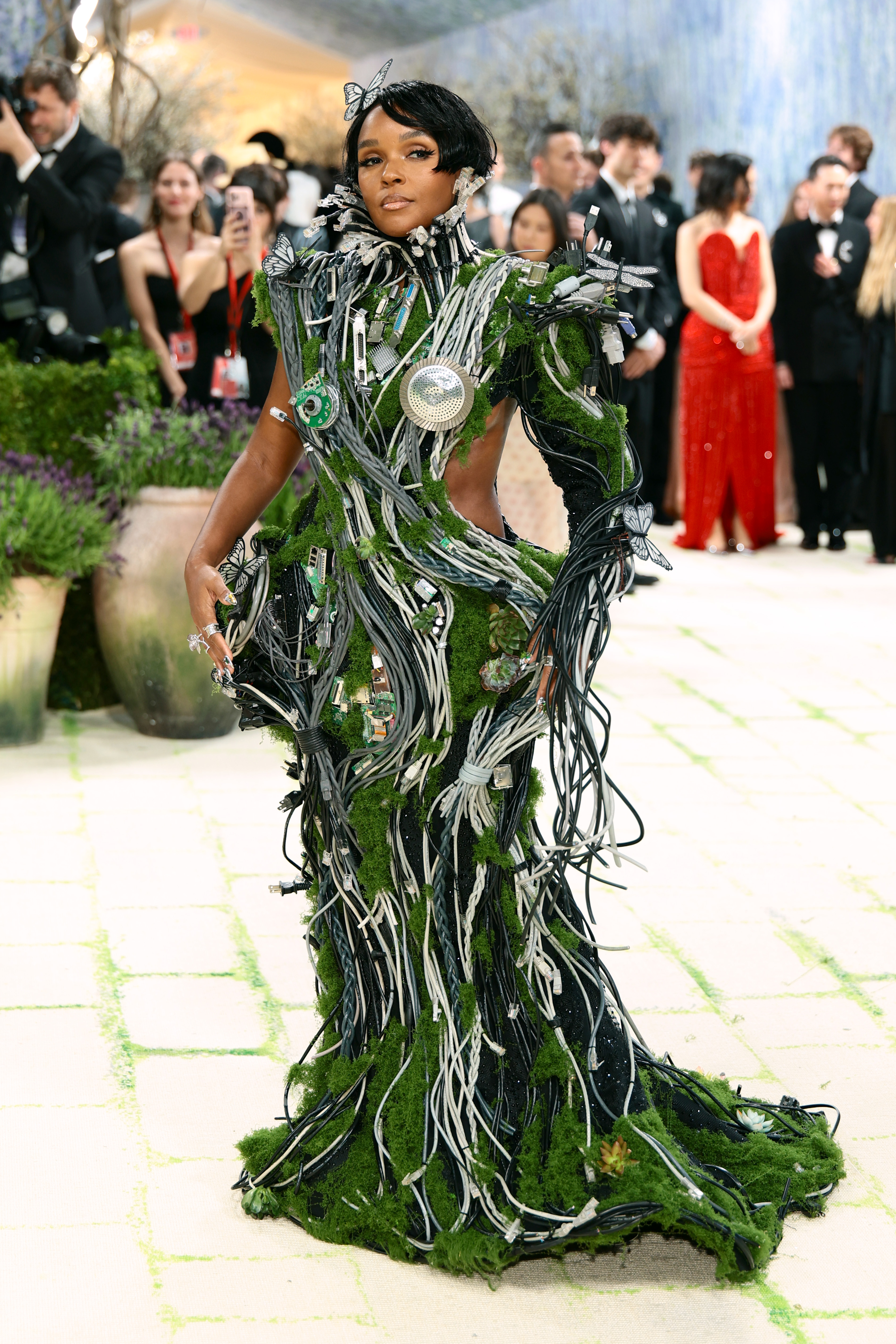 A close front-facing shot of Janelle Monáe's gown revealing its intricate construction: visible circuit boards, USB cables, embedded succulents, a chrome disc centrepiece, and dragonfly and butterfly embellishments nestled amongst moss-covered wiring at the hem.