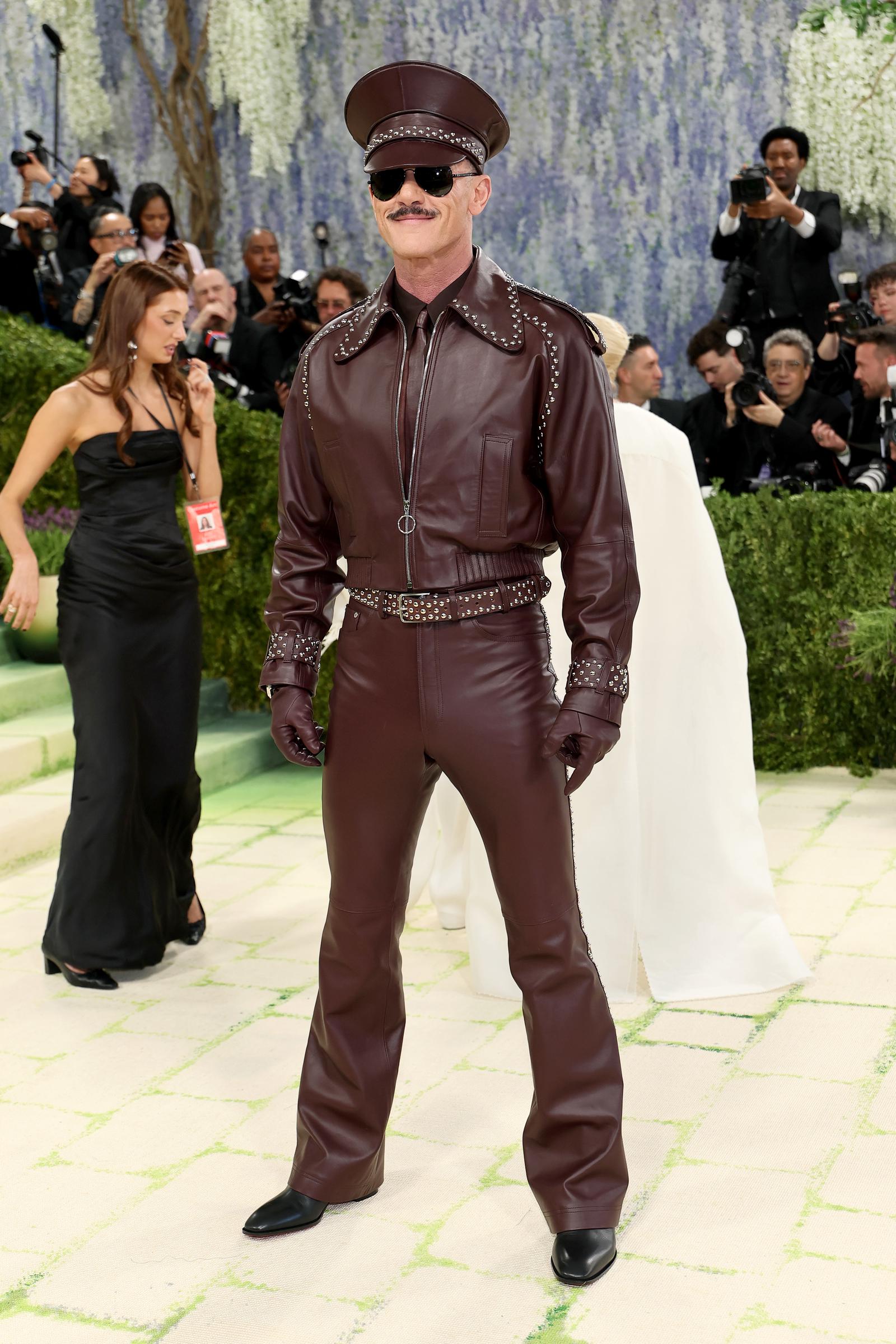 A full-length shot of Luke Evans in his complete oxblood leather look: studded peaked cap, aviator sunglasses, silver-studded collar and cuffs, a double-studded belt, matching leather gloves, flared trousers, and pointed black boots.