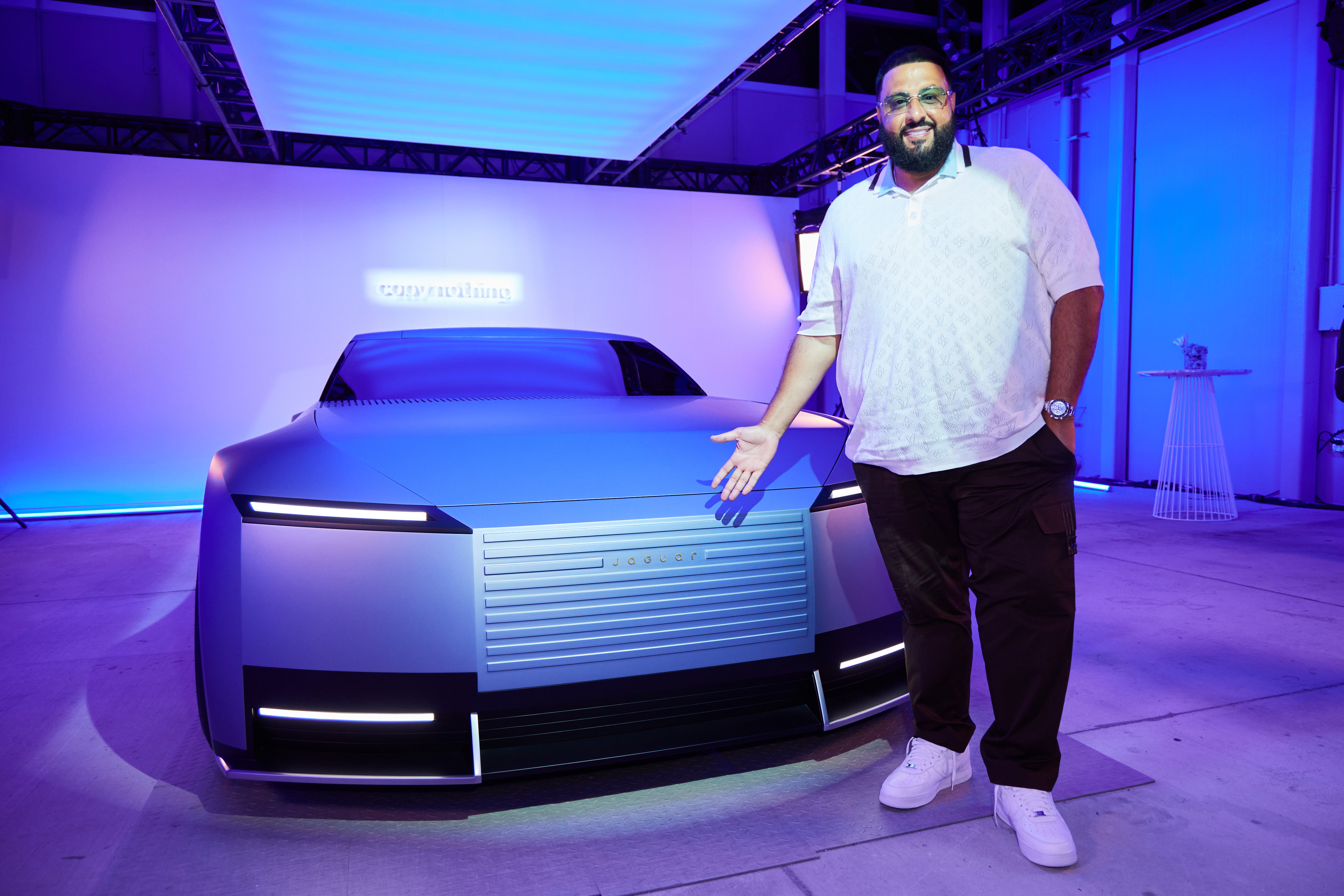 Inside a softly lit Haute Living Celebrating The Future Of Jaguar event, DJ Khaled stands beside a sleek concept vehicle wearing a pale patterned polo, dark trousers, and white sneakers, gesturing toward the futuristic grille as cool blue and violet lighting reflects across the polished surface.