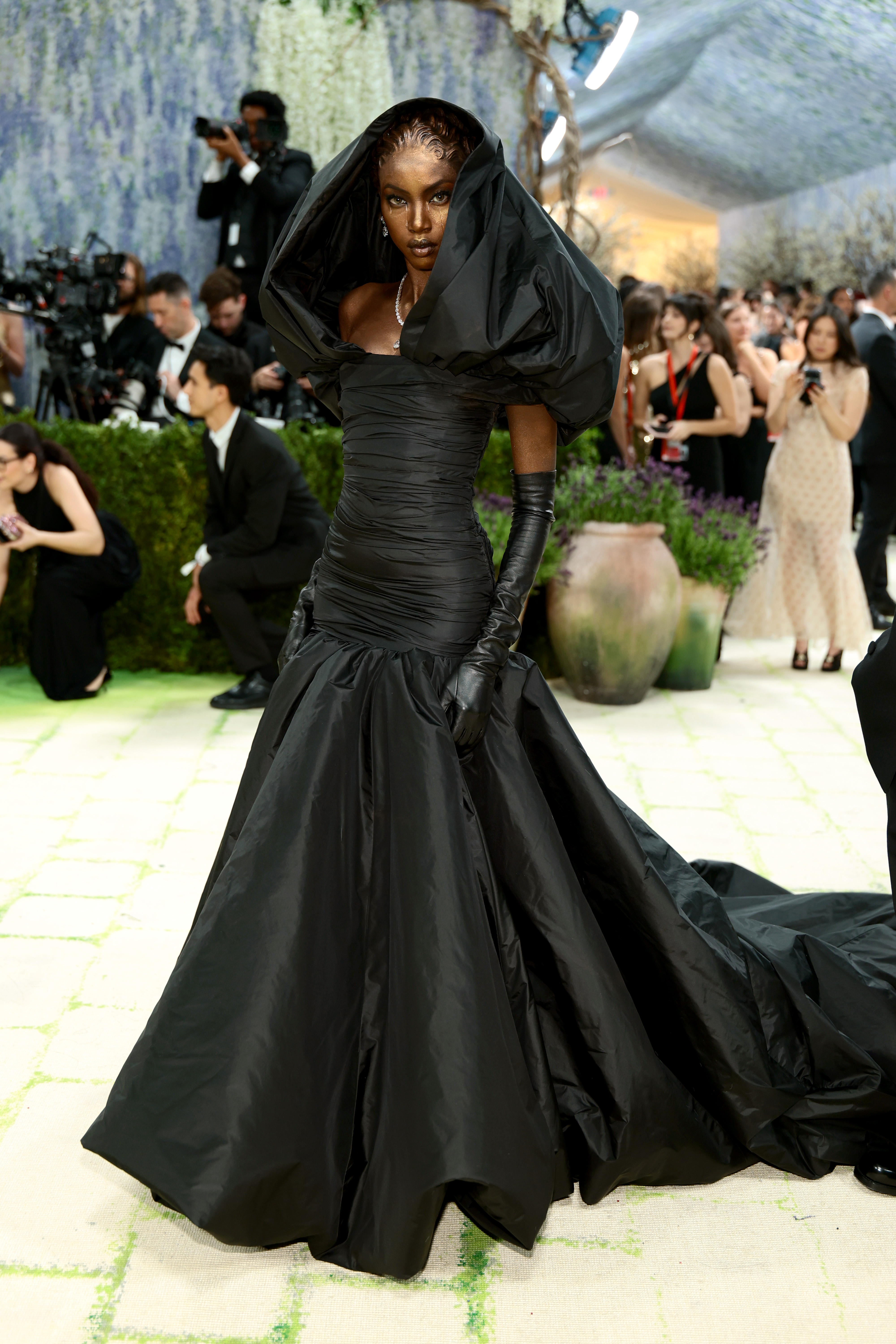 Anok Yai in an all-black ruched strapless ballgown with a voluminous trumpet skirt and sweeping train, a dramatic oversized puffed hood framing her face, long black leather gloves, and a delicate diamond necklace.