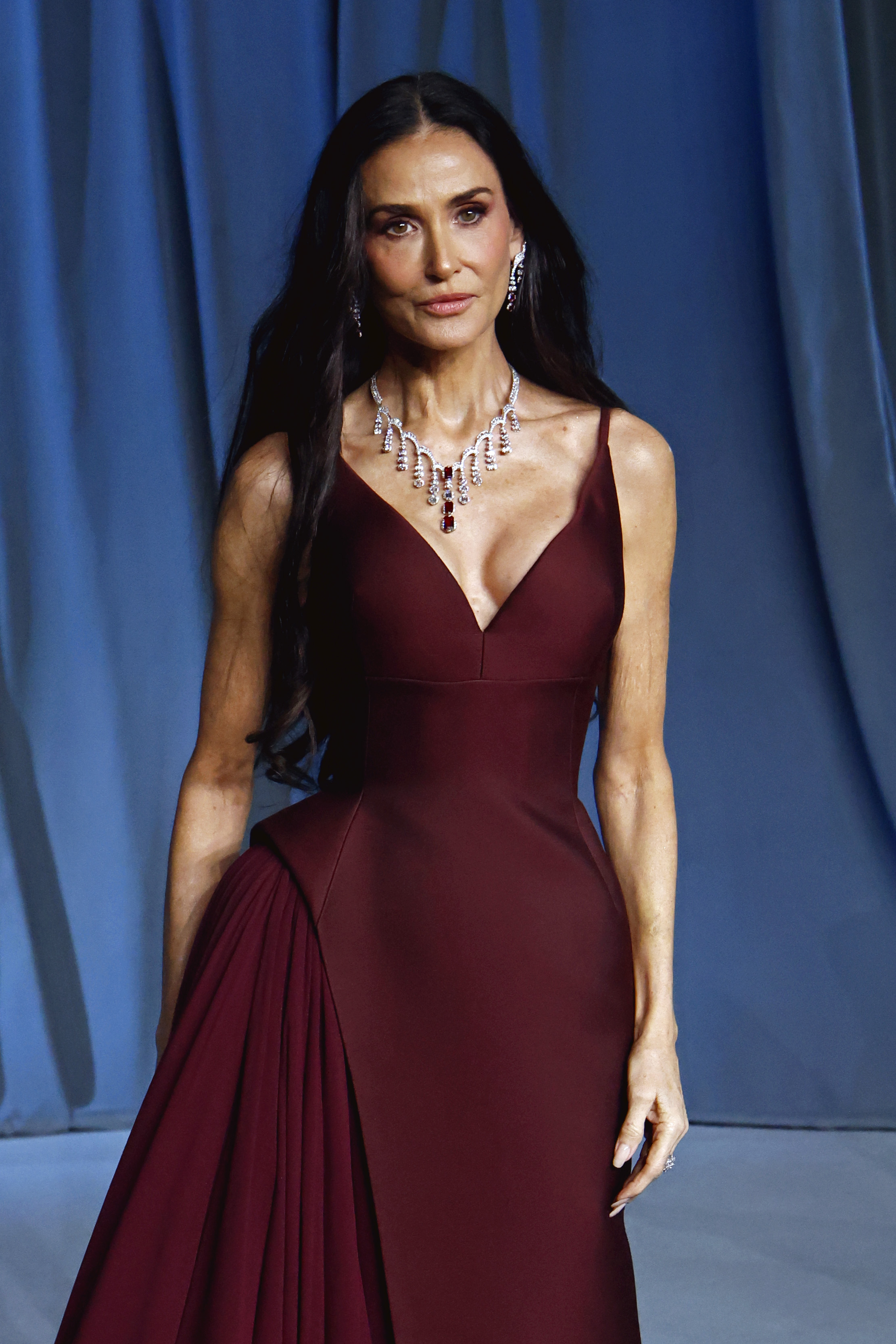 With a fierce, statuesque presence, Demi Moore commands attention in a structured maroon gown, showing off bold pleats and dazzling jewels that radiate Hollywood royalty.