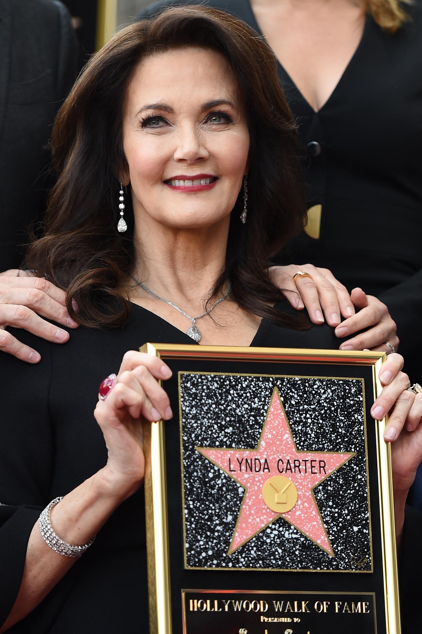 Lynda Carter beams with pride as she holds her Hollywood Walk of Fame plaque during the star ceremony. Dressed in an elegant black ensemble with delicate silver jewelry and drop earrings, she celebrates the career milestone surrounded by loved ones, her signature grace on full display.