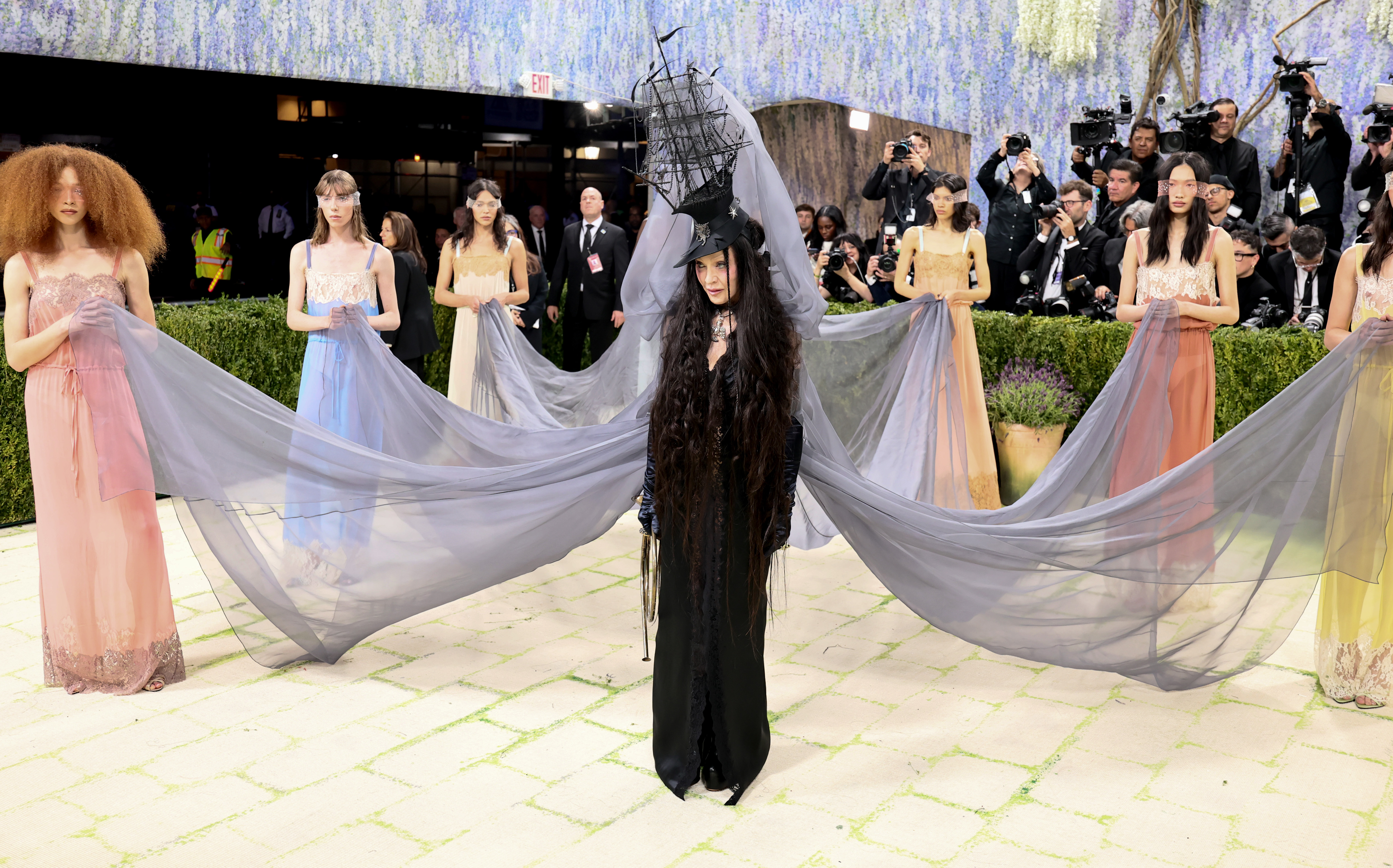 Madonna centred on the carpet flanked by six models in pastel slip dresses — pink, blue, coral and yellow — holding a sweeping grey tulle veil aloft on either side, forming a cathedral-like wingspan across the full width of the frame.