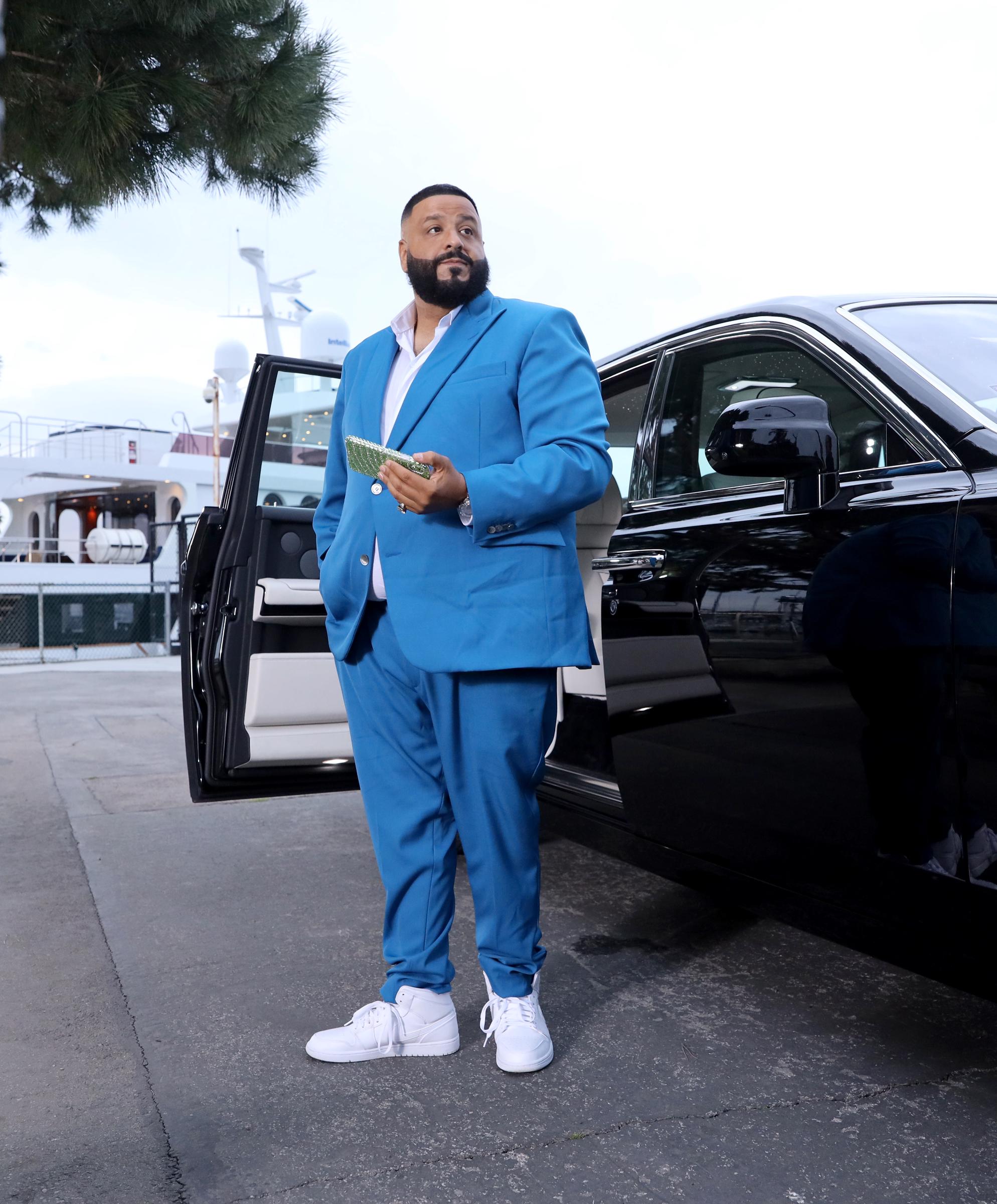 Standing beside an open luxury car door, DJ Khaled is dressed in a vivid blue suit paired with crisp white sneakers, holding a textured clutch as he gazes off-camera with quiet confidence against a marina backdrop that adds a polished, lifestyle-inspired touch to the moment.