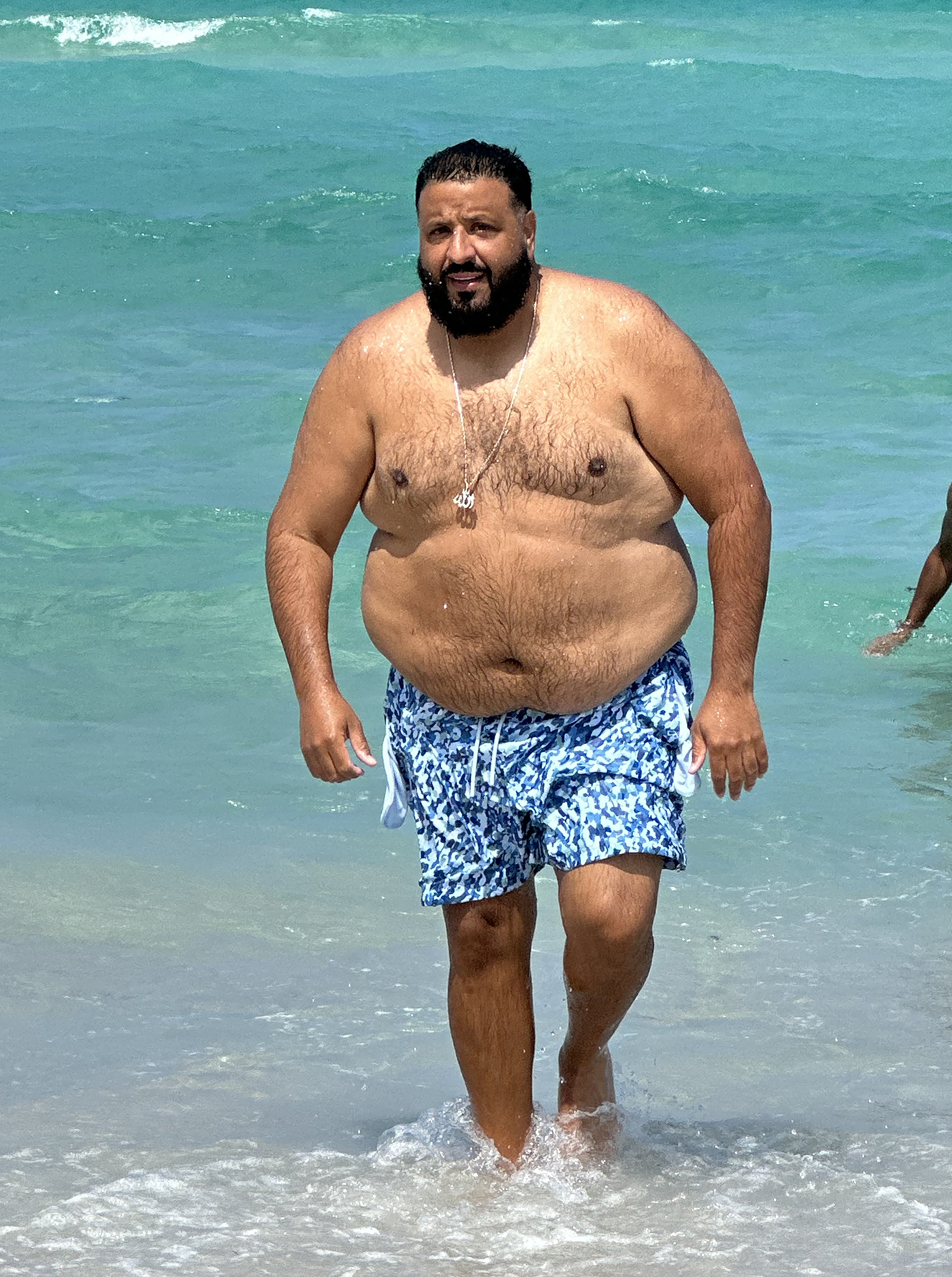 Wading through shallow ocean water, DJ Khaled wears patterned blue swim shorts and a silver chain, his beard damp and hair slicked back as waves ripple behind him under bright daylight.