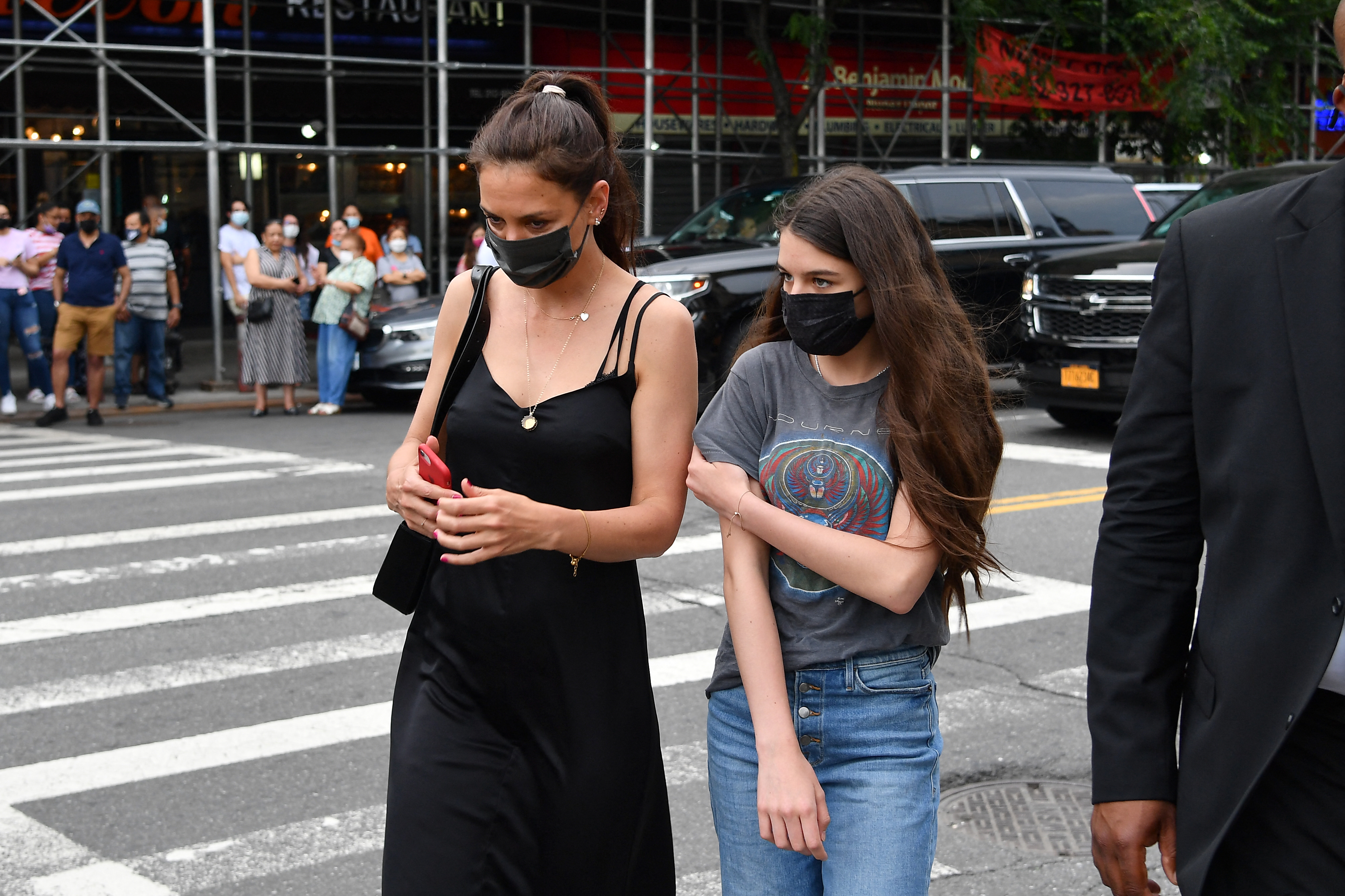 Katie Holmes and Suri walk side-by-side through the city, blending elegance with casual ease. The mom's black dress contrasts beautifully with her daughter's relaxed jeans and band tee, both exuding effortless cool. Their quiet connection amid the urban buzz speaks of love and strength.