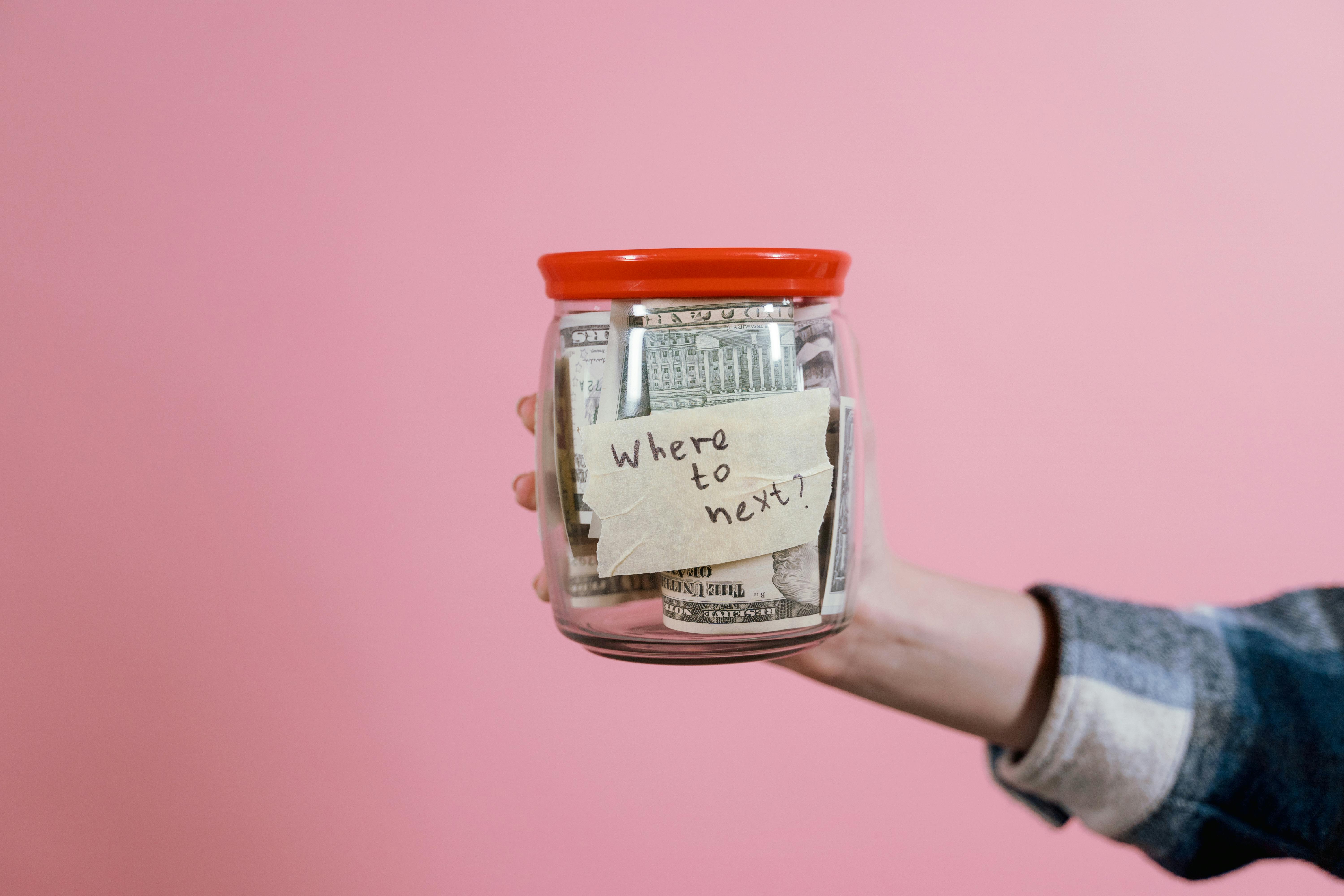 A hand holds a jar full of cash, labeled with a note reading "Where to next?" | Source: Pexels