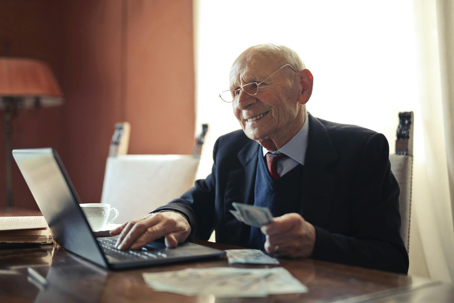 Elderly man holding money in front of a laptop | Source: Pexels