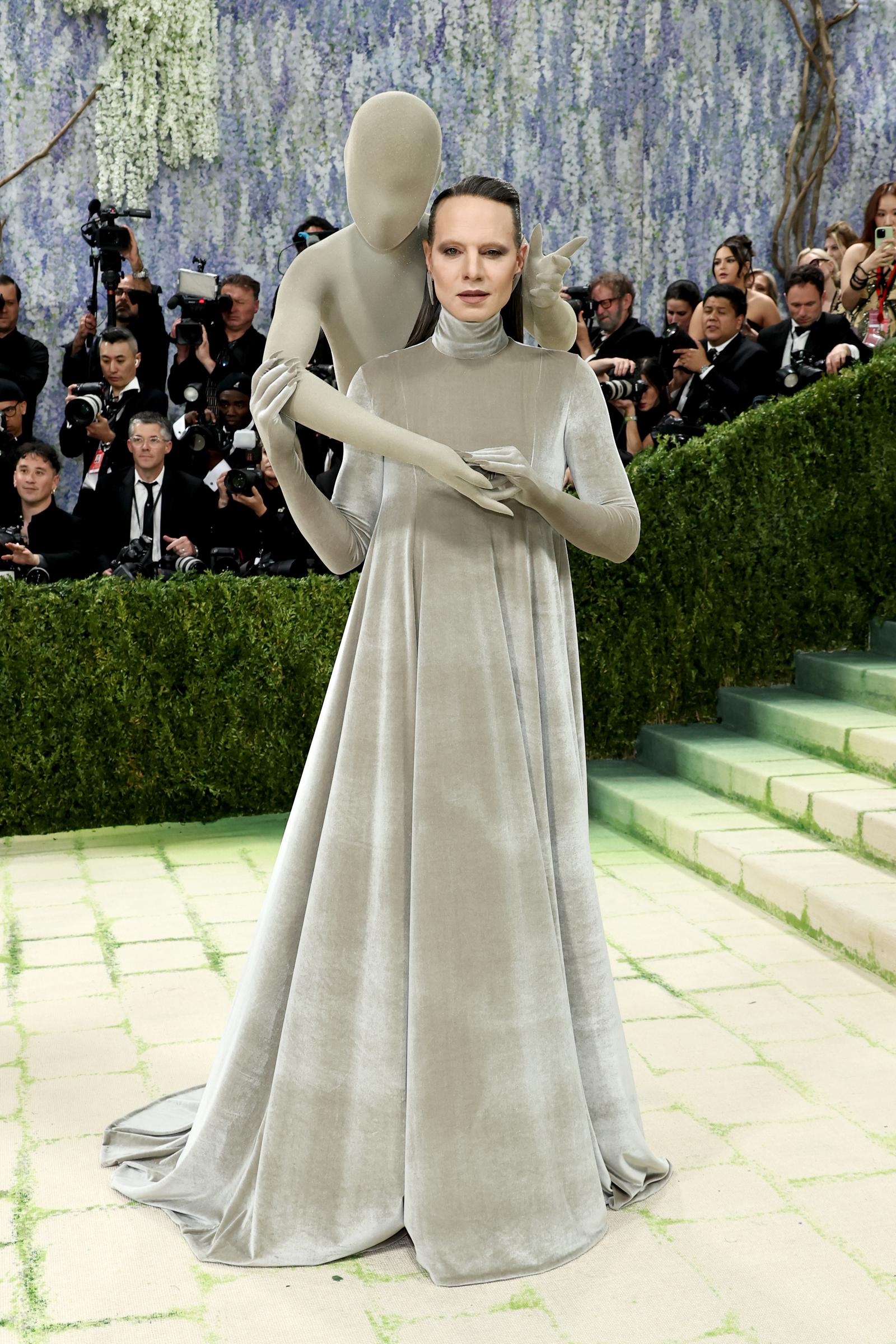 Jordan Roth in a sweeping dove-grey velvet turtleneck gown with matching long gloves, accompanied by a faceless full-body grey mannequin figure that embraces them from behind with outstretched arms.