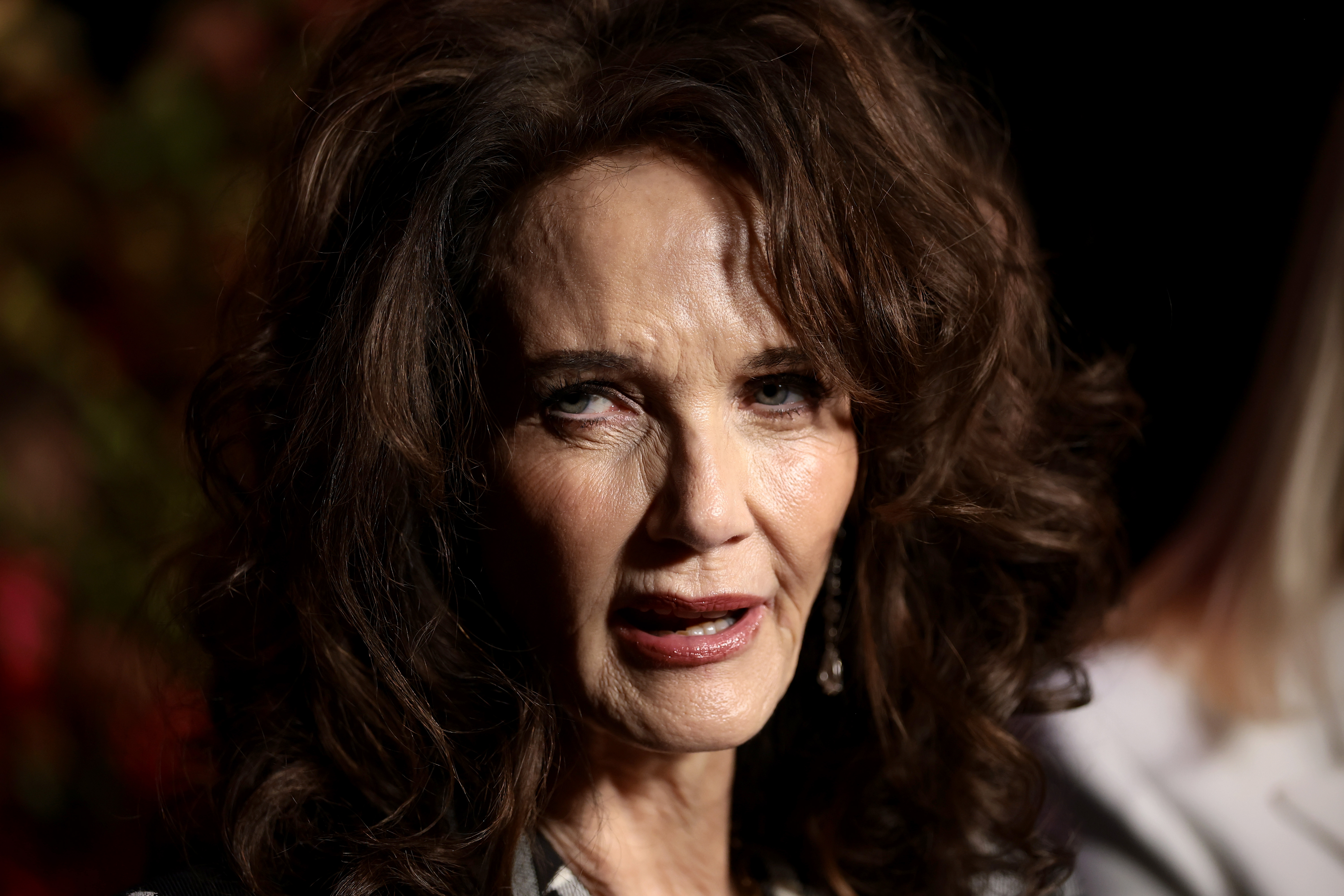Lynda Carter is captured in a dramatic close-up, her expression poised and contemplative under soft, moody lighting. Her voluminous curls frame her face, while subtle makeup and dangling earrings complete the refined, understated look.