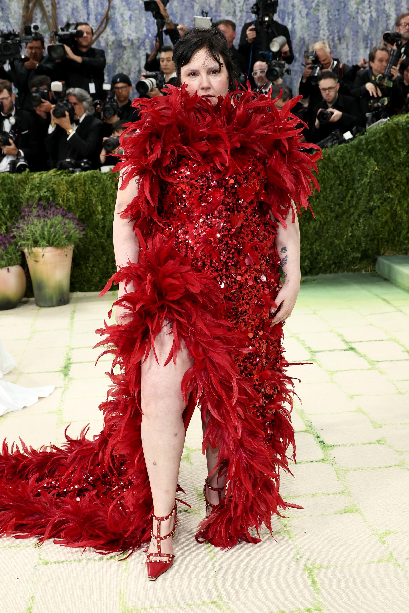 Lena Dunham in a heavily sequinned red gown completely bordered in thick red ostrich feathers from neckline to trailing hem, with studded red T-strap heels and a visible arm tattoo.