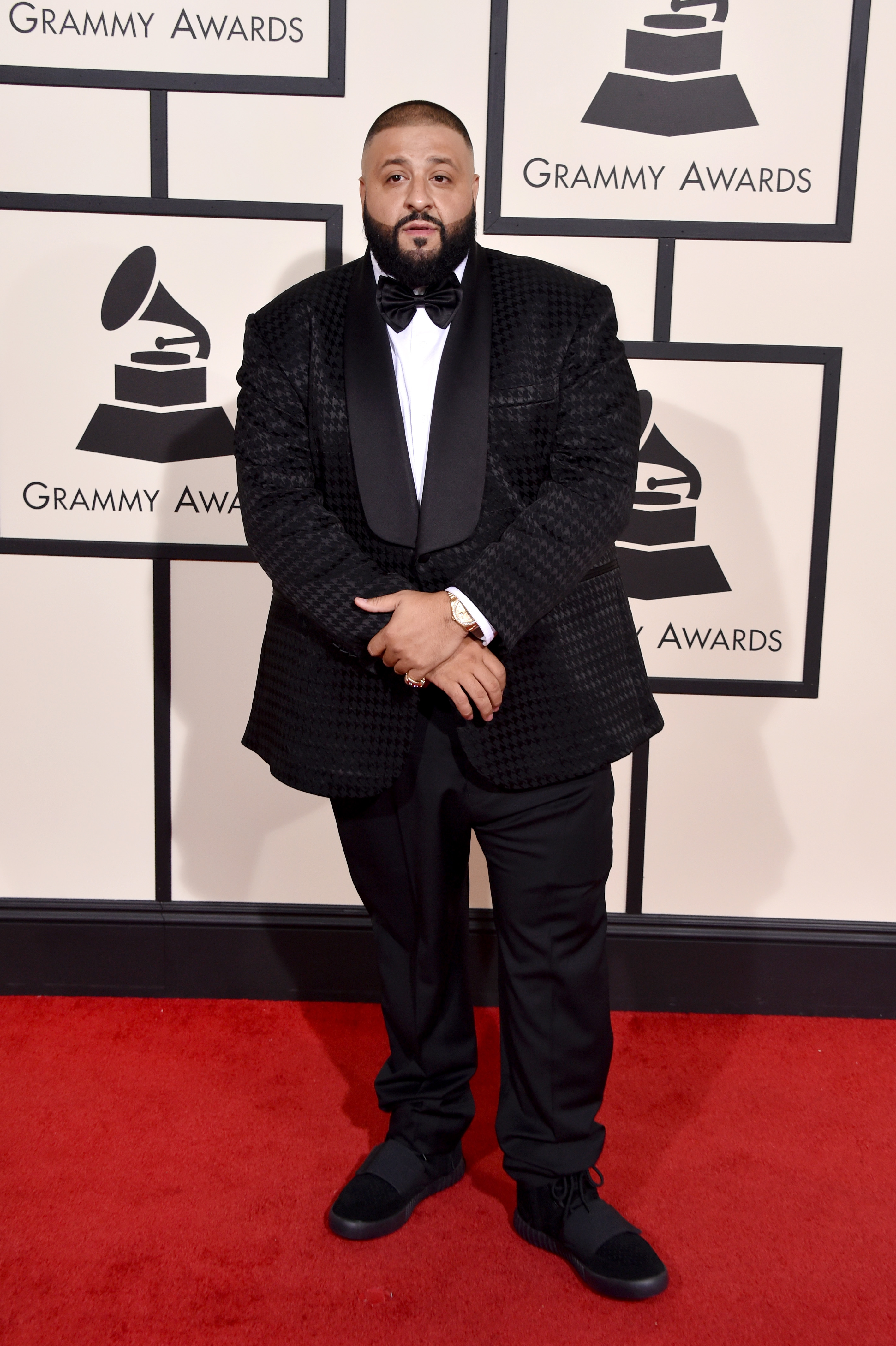At the Grammy Awards, DJ Khaled poses in a textured black tuxedo jacket layered over a crisp white shirt and bow tie, standing against the event’s signature step-and-repeat wall with a composed stance and hands folded neatly in front.