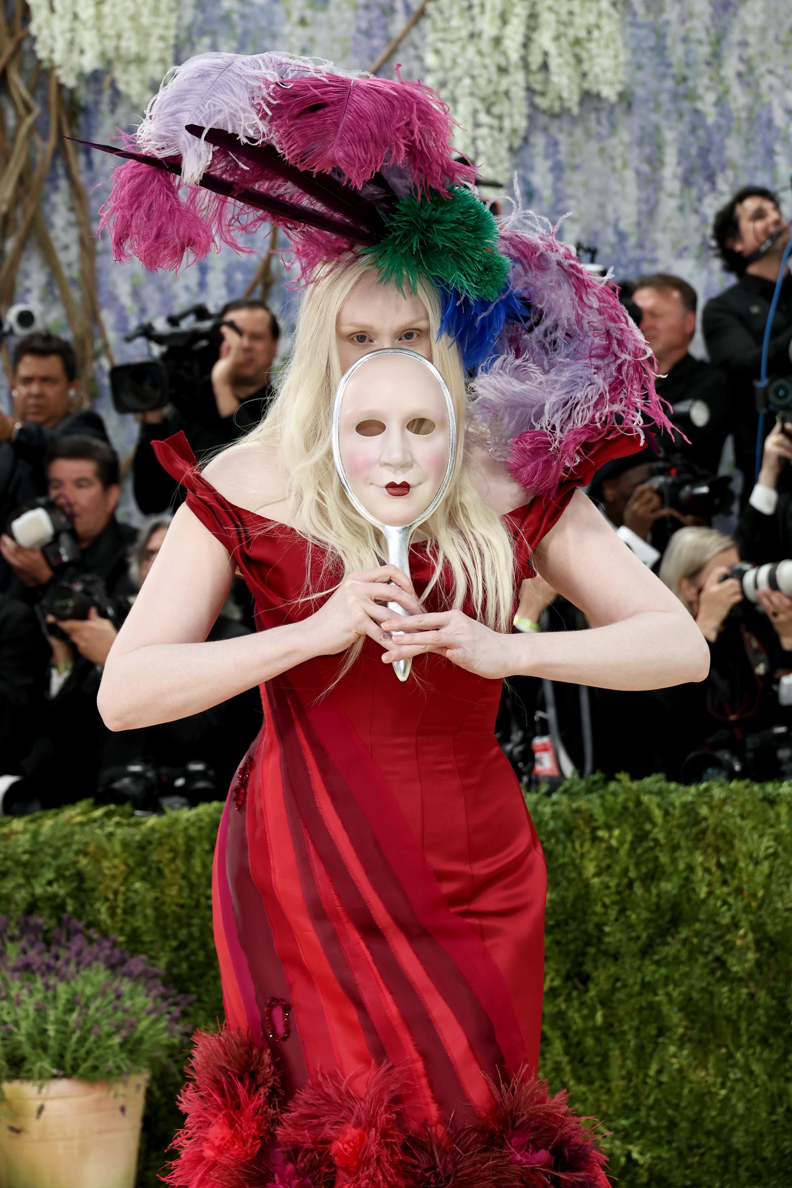 Gwendoline Christie in a red striped off-shoulder gown with a sweeping red feather hem, holding a silver hand-mirror mask painted with red lips up to her face, topped with an explosion of multicoloured ostrich feathers in pink, purple, green and blue.