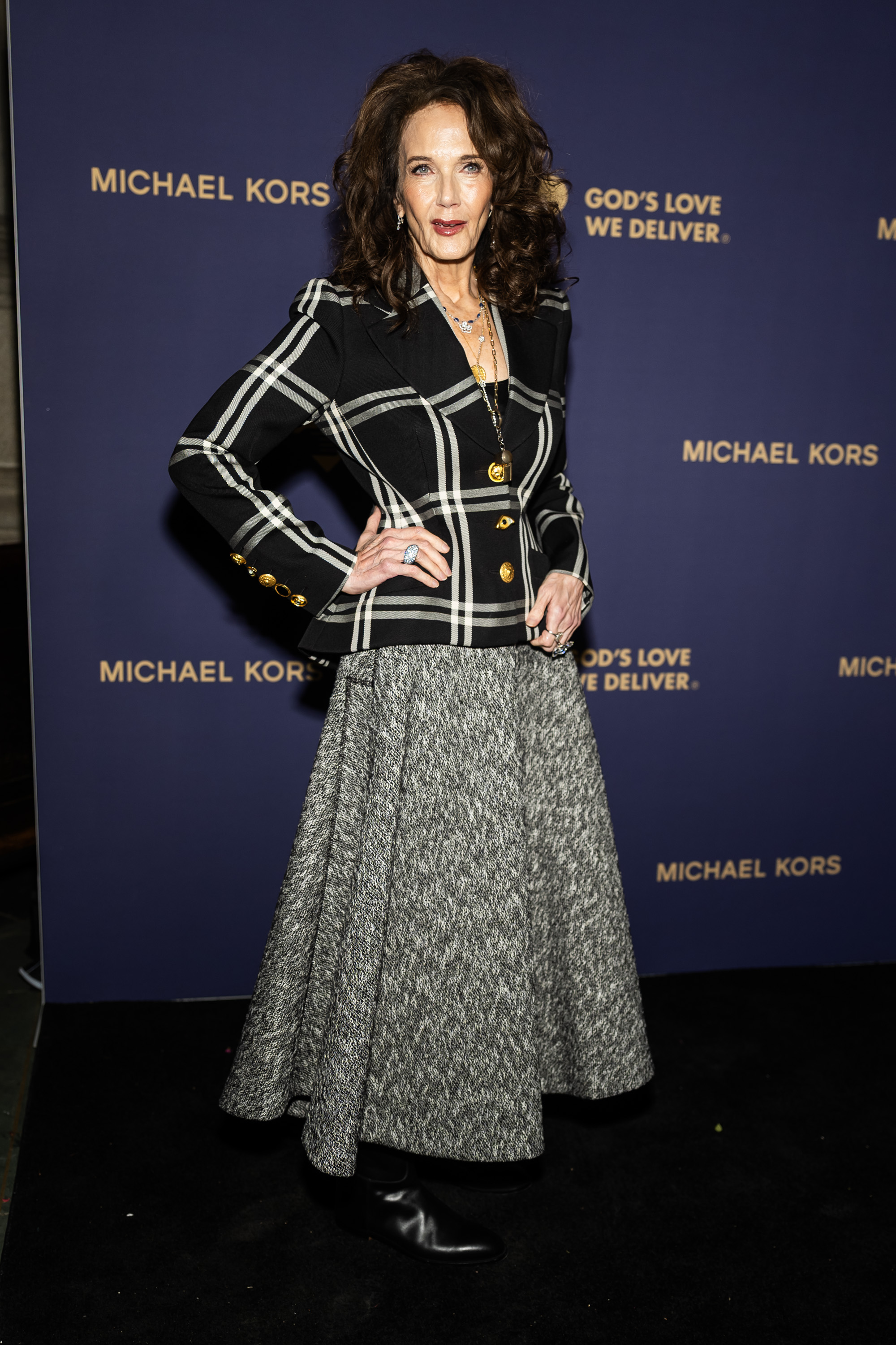 Lynda Carter poses on the red carpet at the God’s Love We Deliver event in a striking black-and-white ensemble. She pairs a tailored plaid blazer with bold gold buttons and a full, textured gray skirt, finishing the look with statement jewelry, leather boots, and voluminous curls.