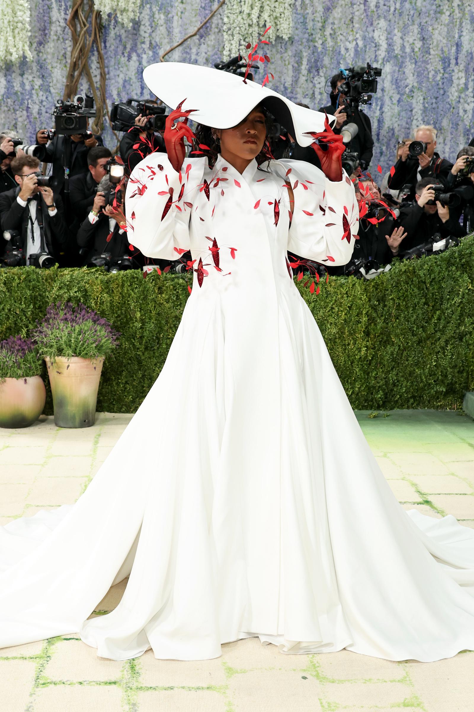 A wider shot of Naomi Osaka showing the full sweep of the white gown's cathedral train, her red-gloved hands raised to the brim of the oversized white hat, with red leaf appliqués scattered across the voluminous structured sleeves and bodice.