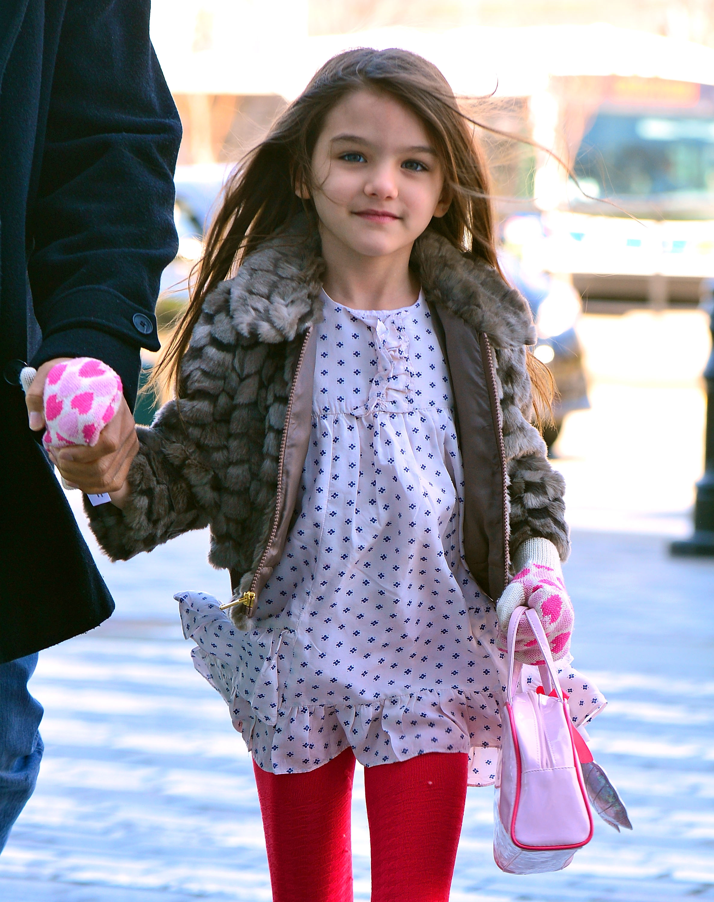 With her hair dancing in the breeze, young Suri walks with poise beyond her years. Dressed in bright tights, a chic coat, and a hint of pink flair, she exudes effortless charm. Her soft smile captures innocence wrapped in budding confidence.