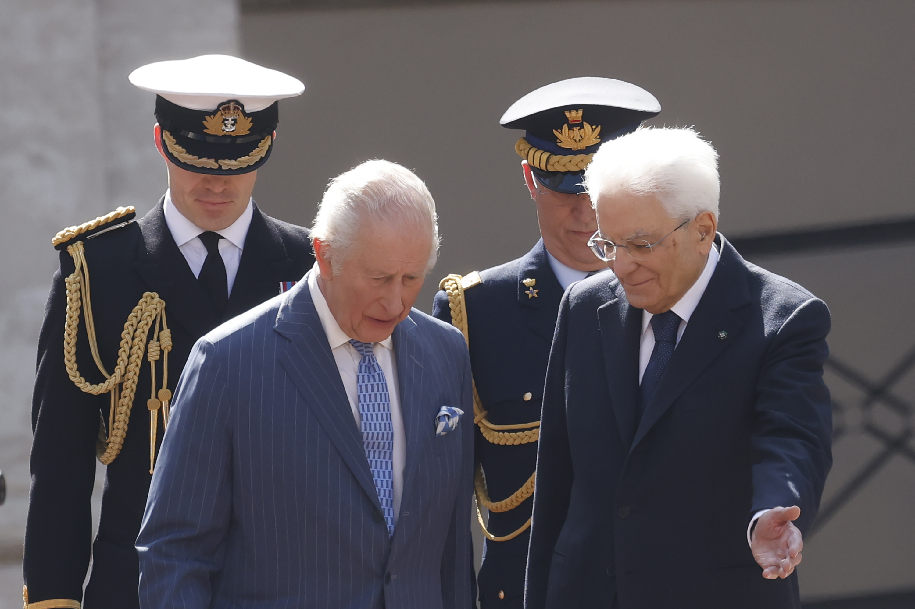 President Mattarella gestures graciously as he walks alongside King Charles III, guiding him during their official meeting at the Quirinale Palace. Dressed in coordinated formal attire, the two heads of state appear deep in discussion, trailed by decorated military personnel.