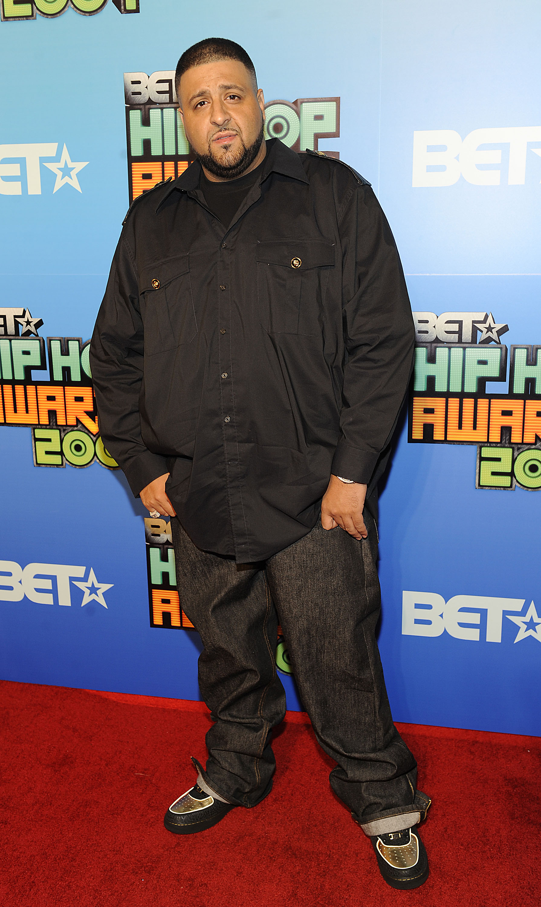 At the BET Hip Hop Awards, DJ Khaled steps out in a black button-up shirt with front pockets and relaxed-fit jeans, completing the look with low-top sneakers as he stands against the blue and orange event backdrop.