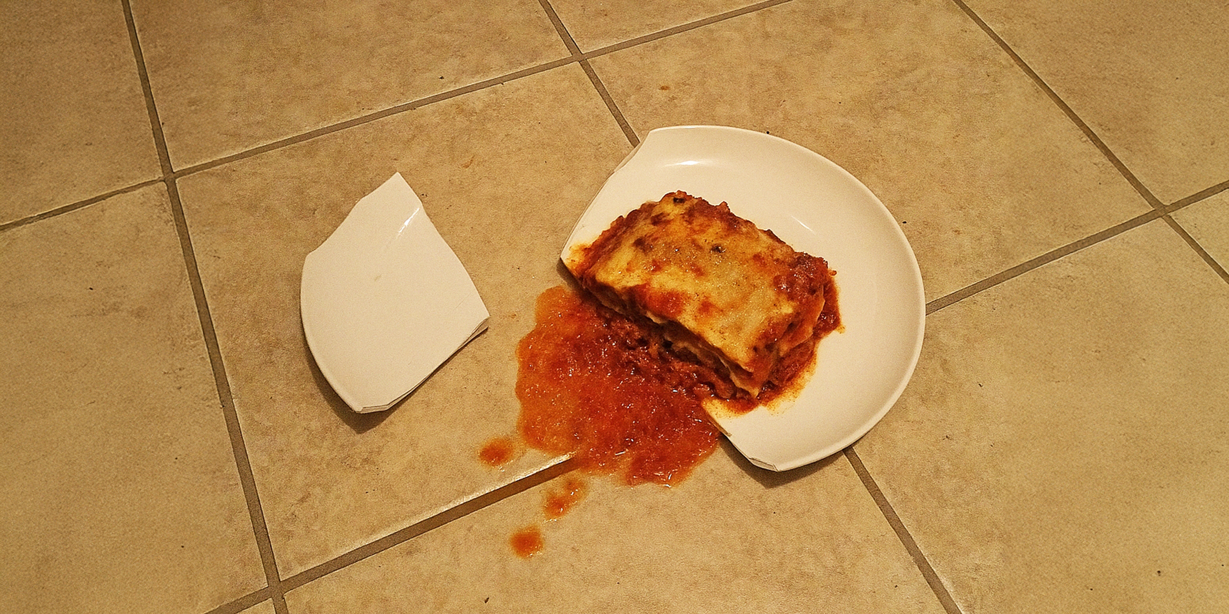 A broken plate lying on a tiled floor, with a serving of lasagna spilled and smeared beside it | Source: The Celebritist