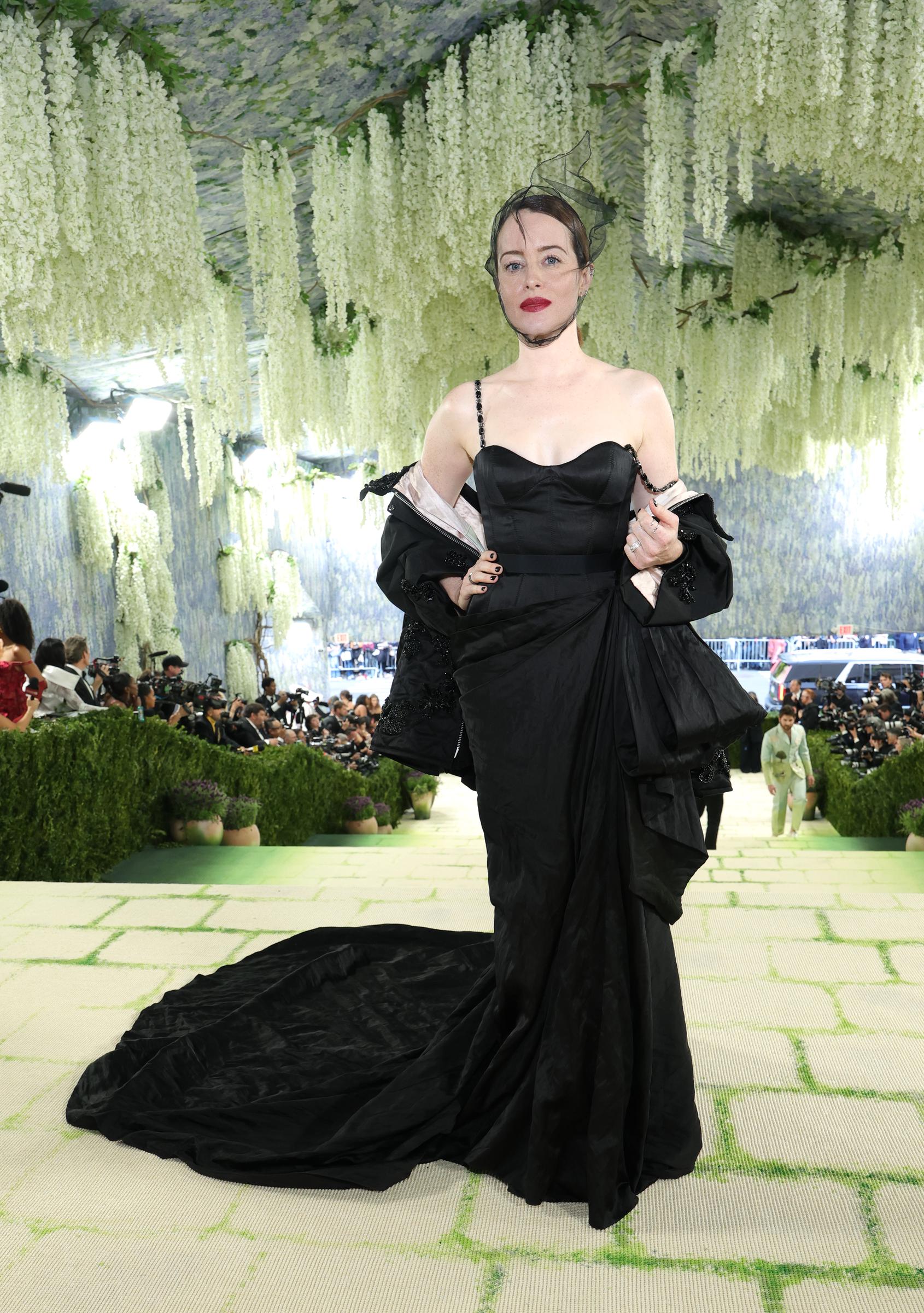Claire Foy in a black sweetheart-neckline satin gown with a long train, a draped off-shoulder jacket with beaded trim, a delicate black neck choker, small black net fascinator, red lips and dark nails.