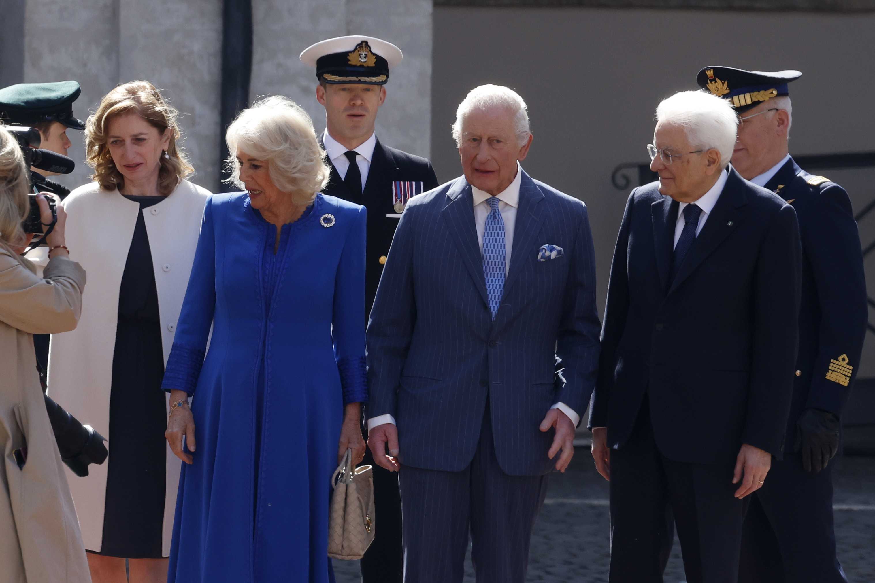 In a formal moment outside the Quirinale Palace, Charles and Camilla are welcomed by Italian President Sergio Mattarella and his daughter. Both monarch and president appear engaged in conversation. Uniformed officers stand at attention behind them, adding to the atmosphere of diplomatic ceremony.