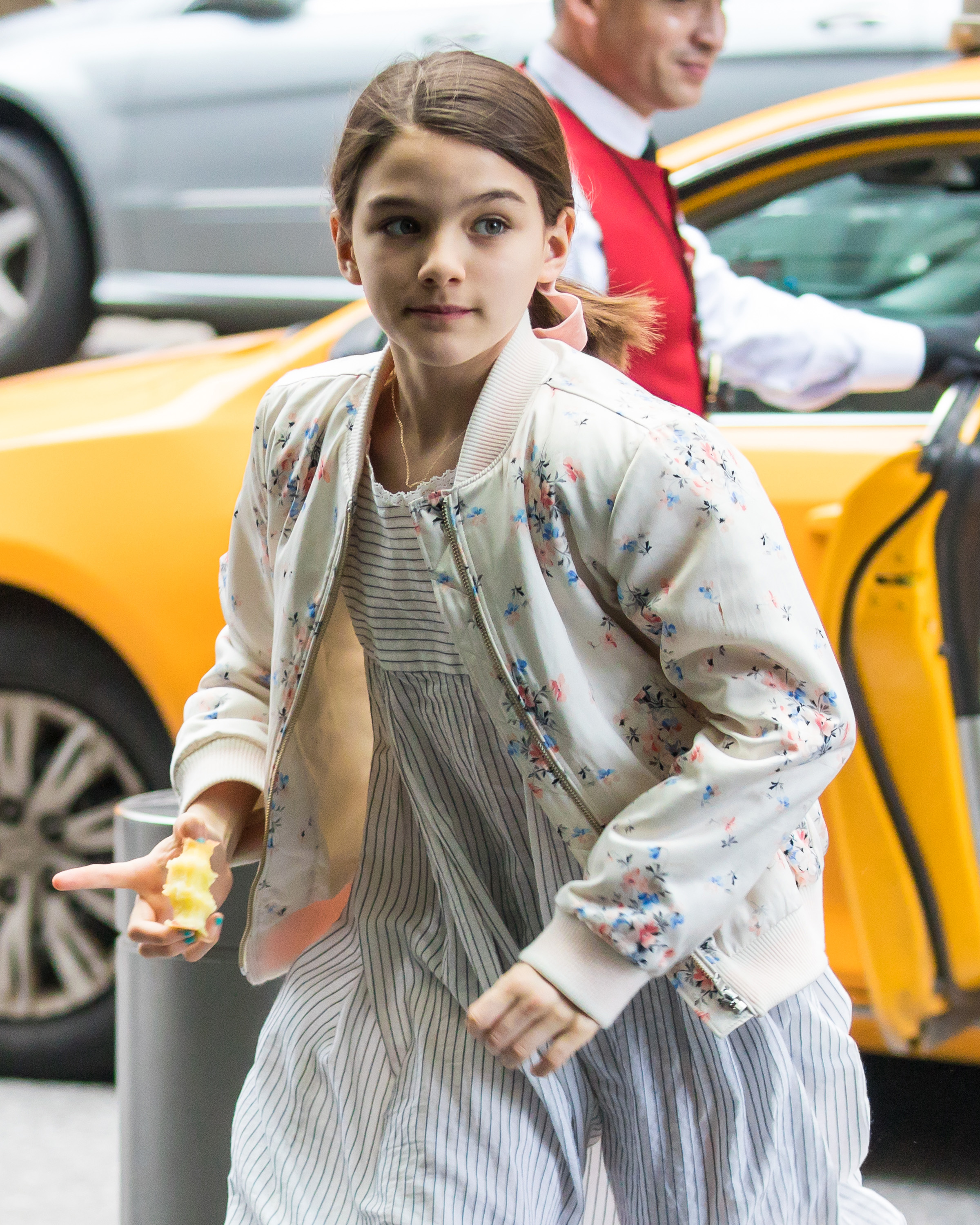 Stepping out of a taxi with poise, Suri Cruise carries a quiet confidence beyond her years. Dressed in a soft floral jacket and striped dress, she embodies youthful grace and ease. With an apple in hand and curiosity in her eyes, she captures the essence of effortless charm.