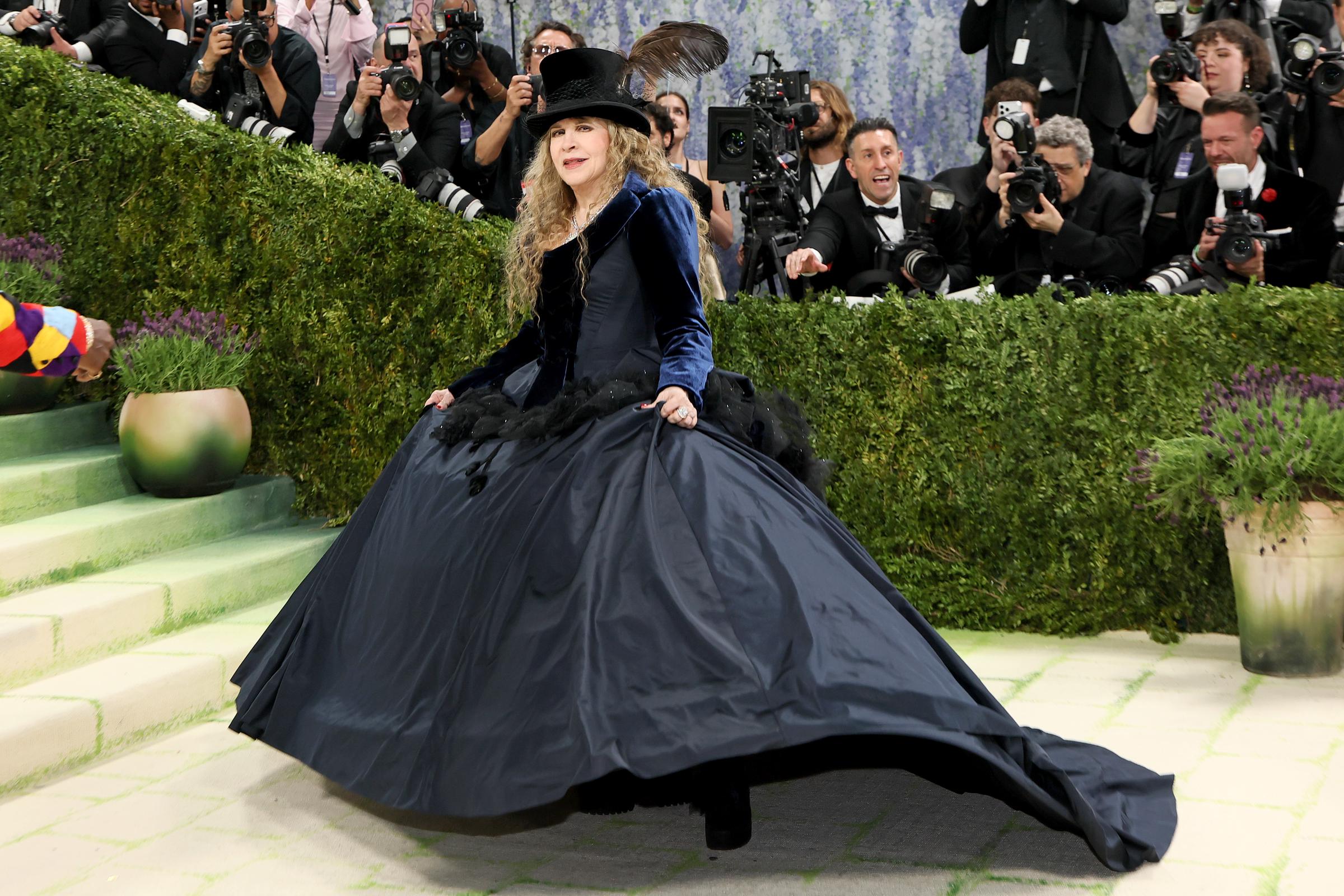 Stevie Nicks caught mid-twirl on the carpet, her sweeping navy taffeta ballgown billowing dramatically outward, paired with a deep blue velvet jacket, black feather-trimmed hem, and a black top hat adorned with a single dark feather.