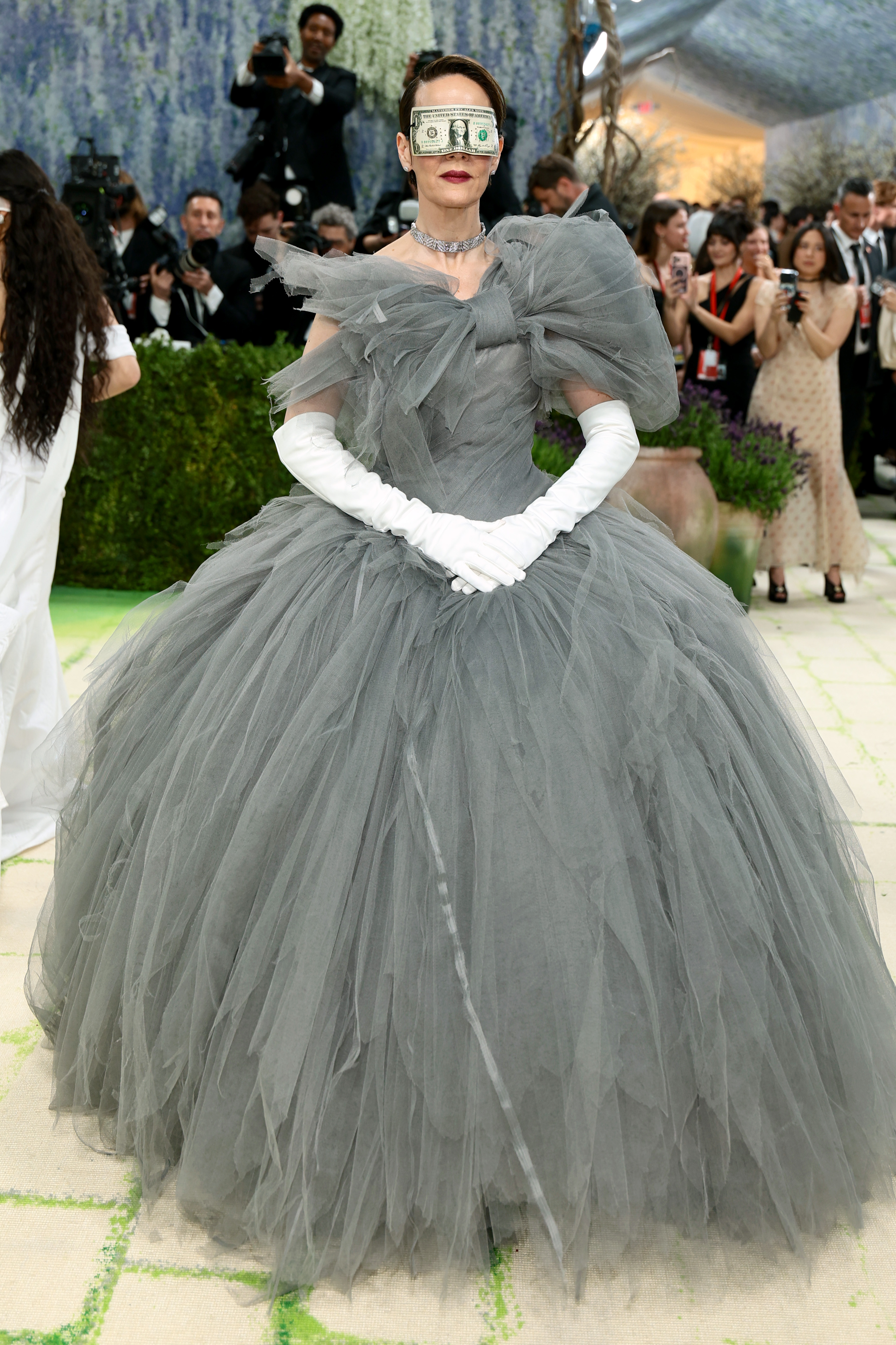 Sarah Paulson in an enormous deconstructed grey tulle ballgown with a ruffled off-shoulder bodice, white opera gloves and a diamond choker, with a US dollar bill worn as a blindfold across her eyes.