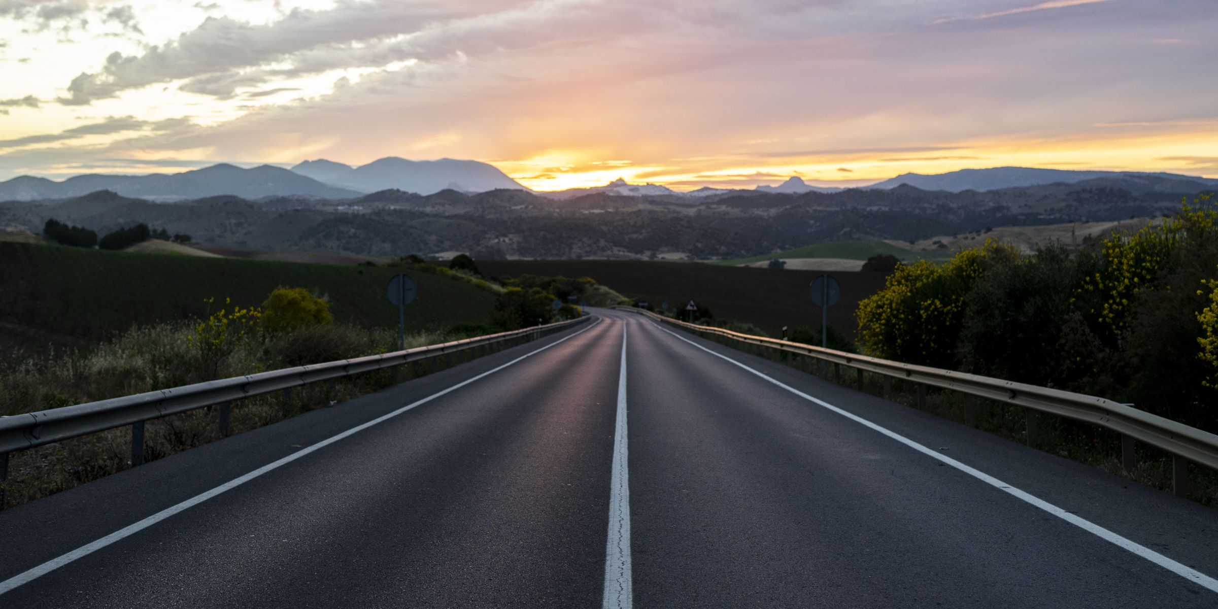 A view of an empty highway surrounded by hills under a cloudy sunset | Source: No source