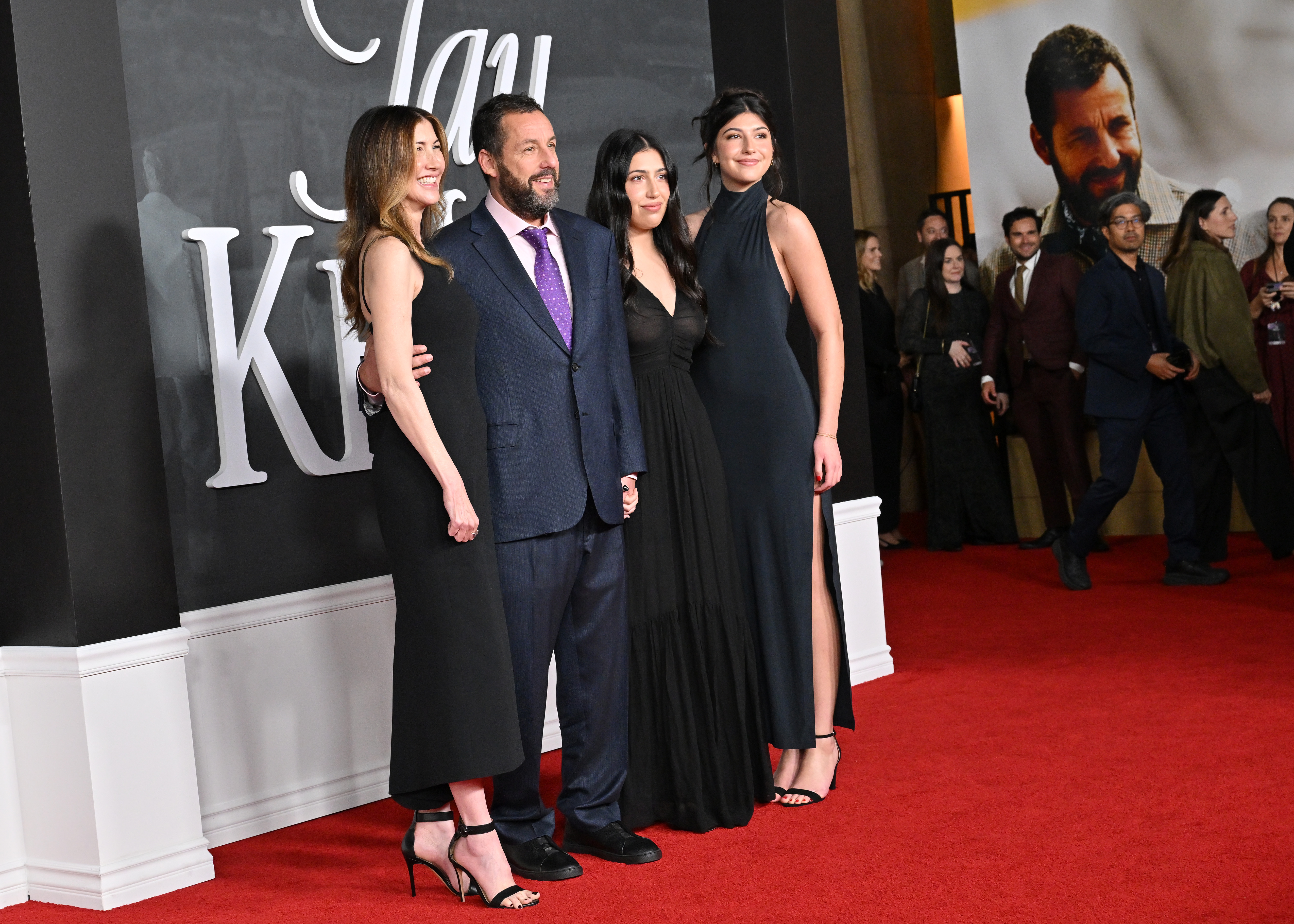 Jackie, Adam, Sadie, and Sunny Sandler look like the perfect celebrity family as they pose for photos at the "Jay Kelly" premiere. This shot stands out even more, capturing a movie poster featuring Adam on the cover in the background.