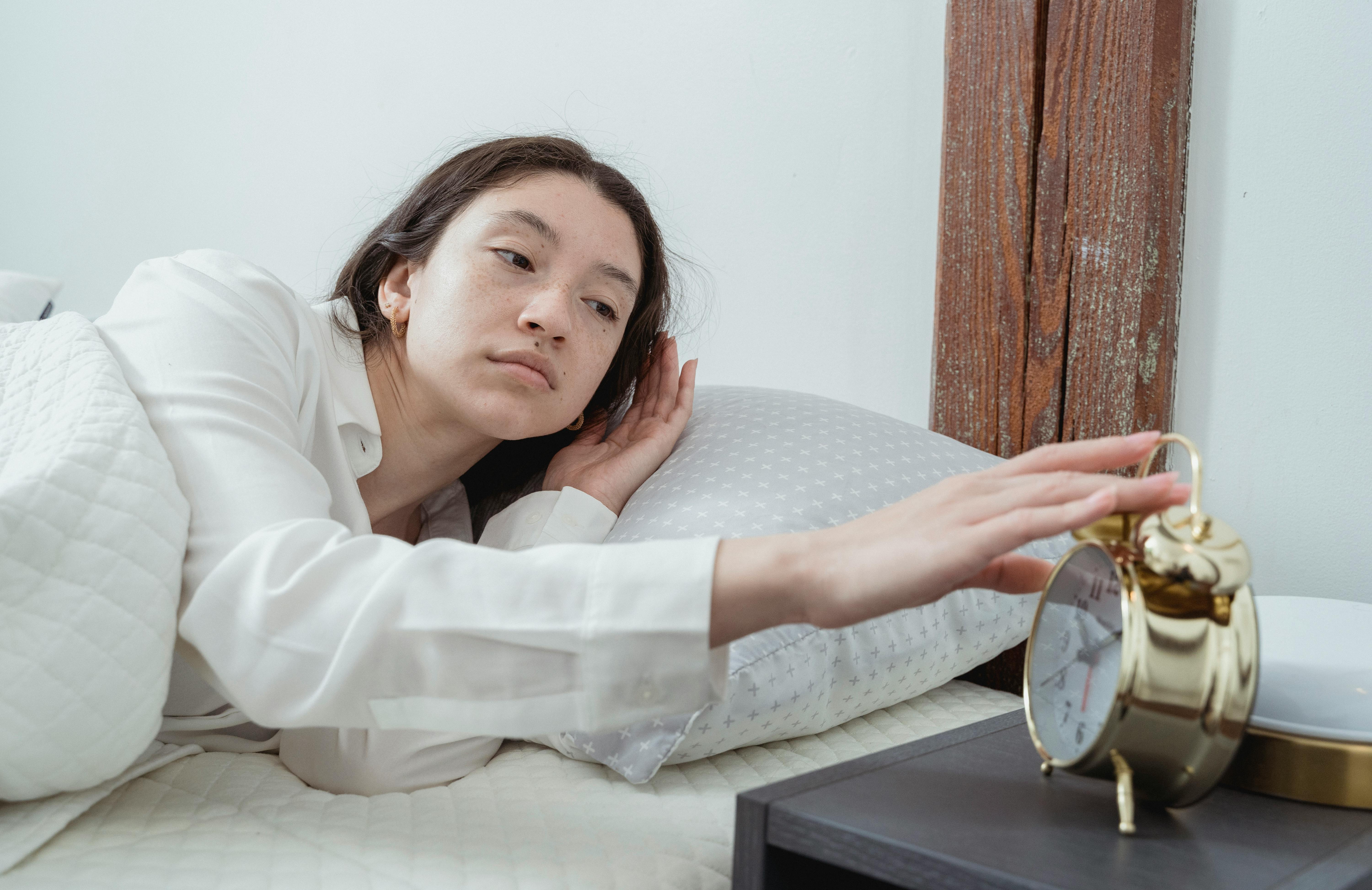 A woman looks tired as she reaches to turn off a gold alarm clock while lying in bed. | Source: Pexels