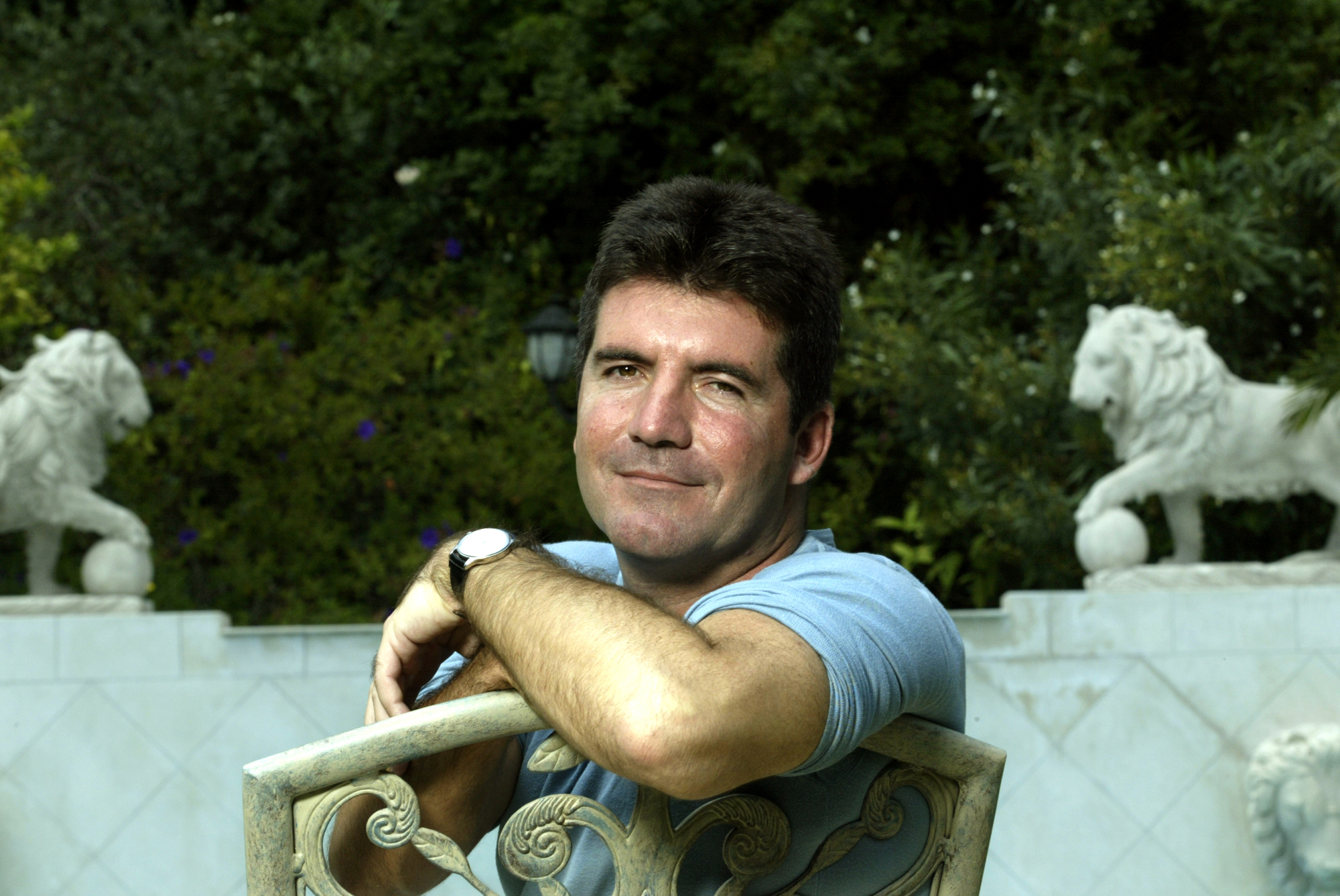 In June 2003, a rare glimpse behind the curtain — Cowell leans pensively against a garden chair at a private Beverly Hills estate. The lush backdrop and classical statues offer a stark contrast to the storm of showbiz soon to come.