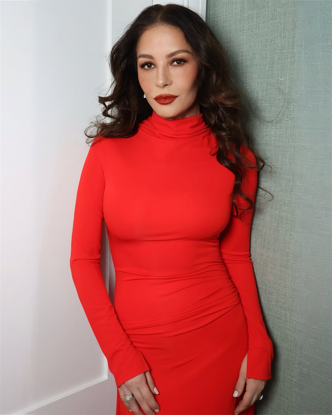 Her 2025 appearance blends classic glamour with modern flair as Zeta-Jones opts for a bright red, curve-hugging turtleneck dress, complemented by brown waves and bold makeup.